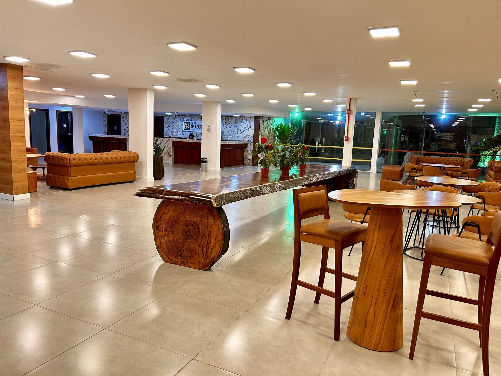 Hall de entrada com assentos, mesa grande de madeira, banquetas e balcão de recepção. Cores neutras e iluminação geral.