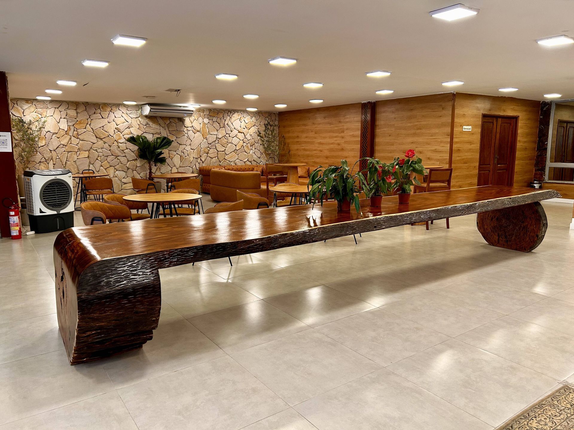 Grande mesa de madeira em uma área de espera com outros assentos, uma parede de tijolos e vasos de plantas.