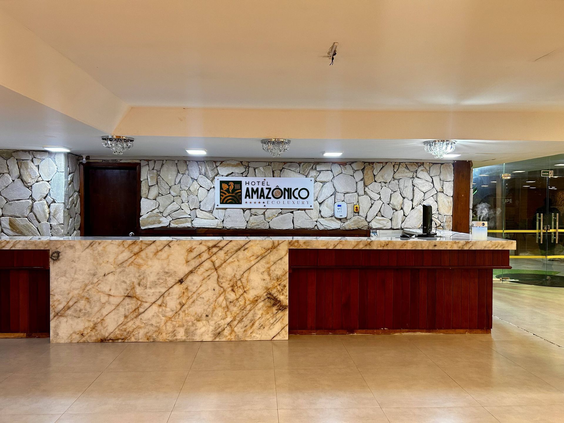 Balcão de recepção do hotel com detalhes em mármore e madeira, paredes bege e placa