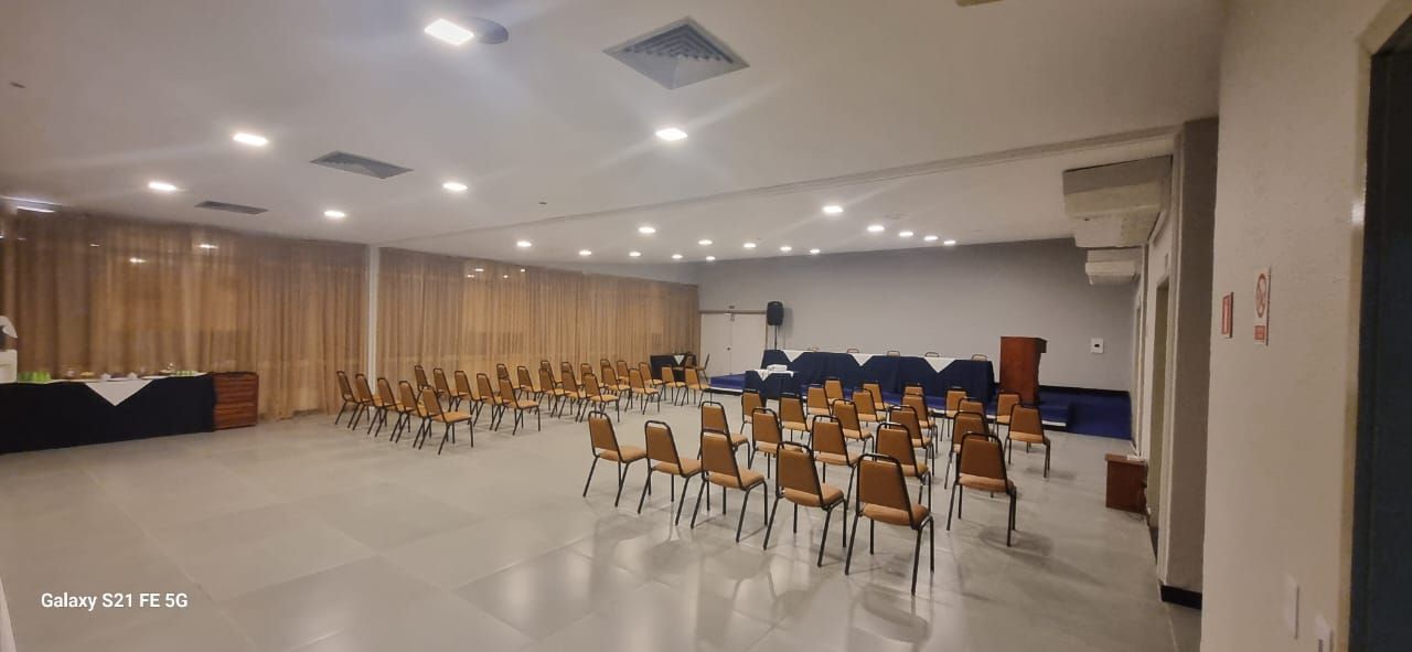 Sala de conferências vazia com fileiras de cadeiras, painel de madeira ao fundo e luzes brilhantes no teto.