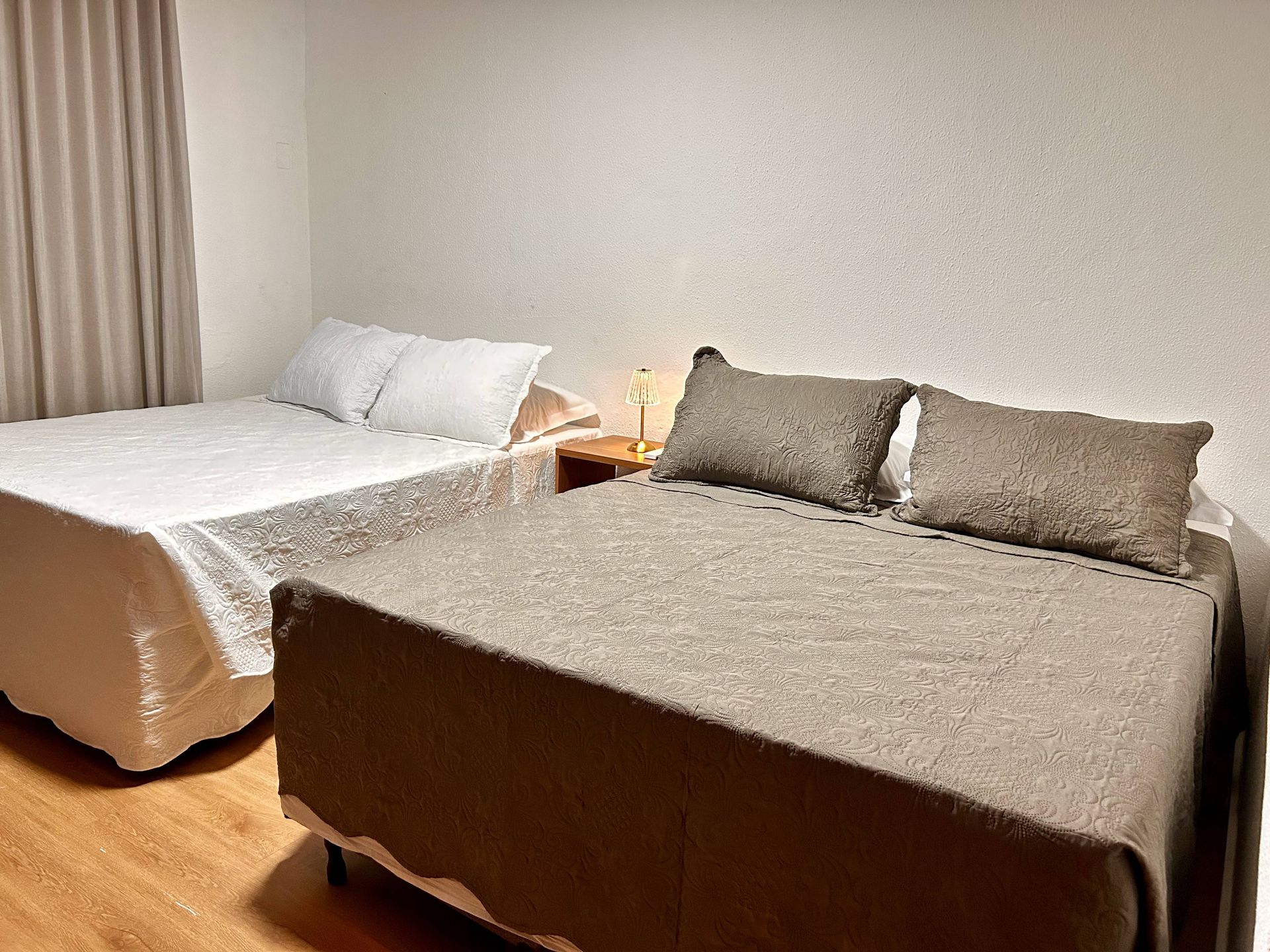 Duas camas em um quarto; uma com roupa de cama branca, a outra com roupa de cama marrom. Pequena mesa de cabeceira com abajur entre elas.