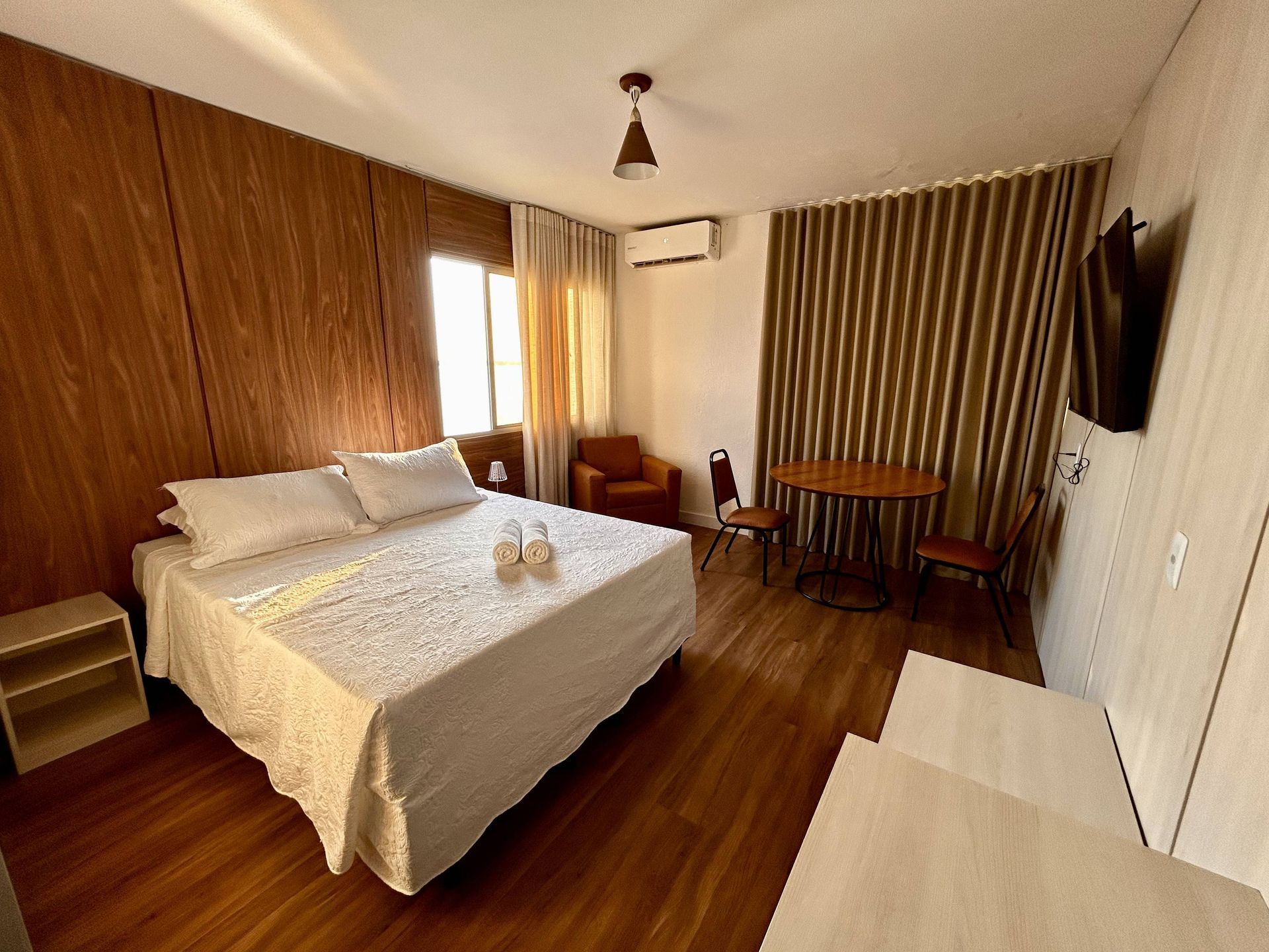 Quarto com painéis e piso de madeira, cama, mesinha com cadeiras, janela, TV e poltrona.