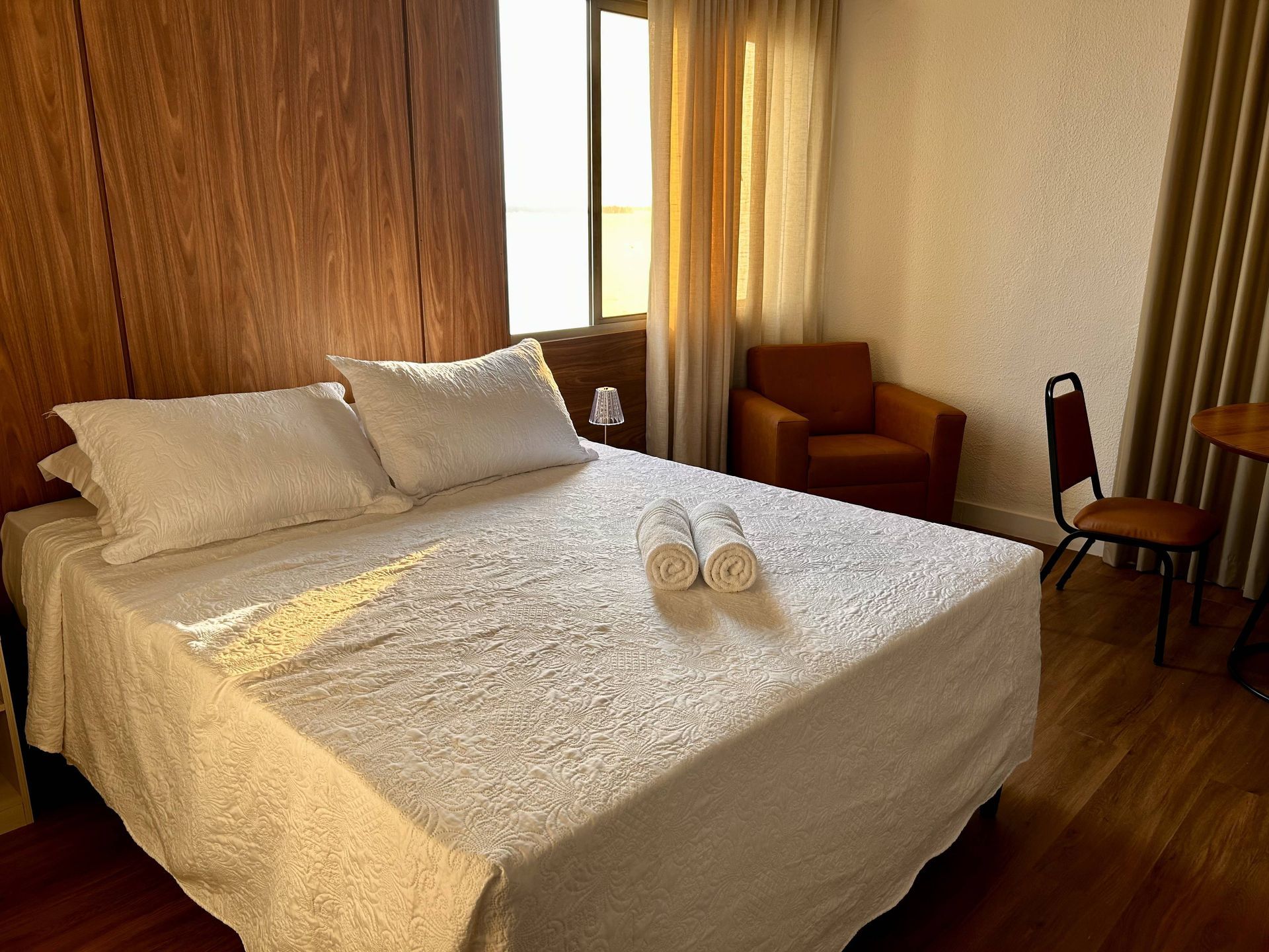 Quarto com cama, toalhas, cadeira e poltrona perto de uma janela com cortinas.