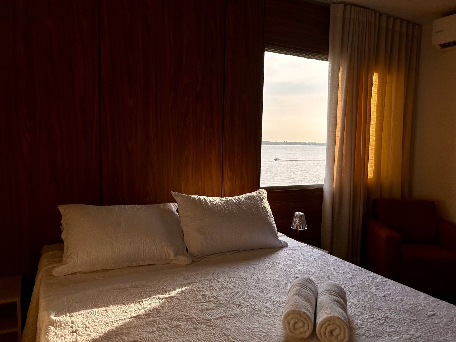 Quarto com cama, travesseiros, toalhas e janela com vista para o mar.