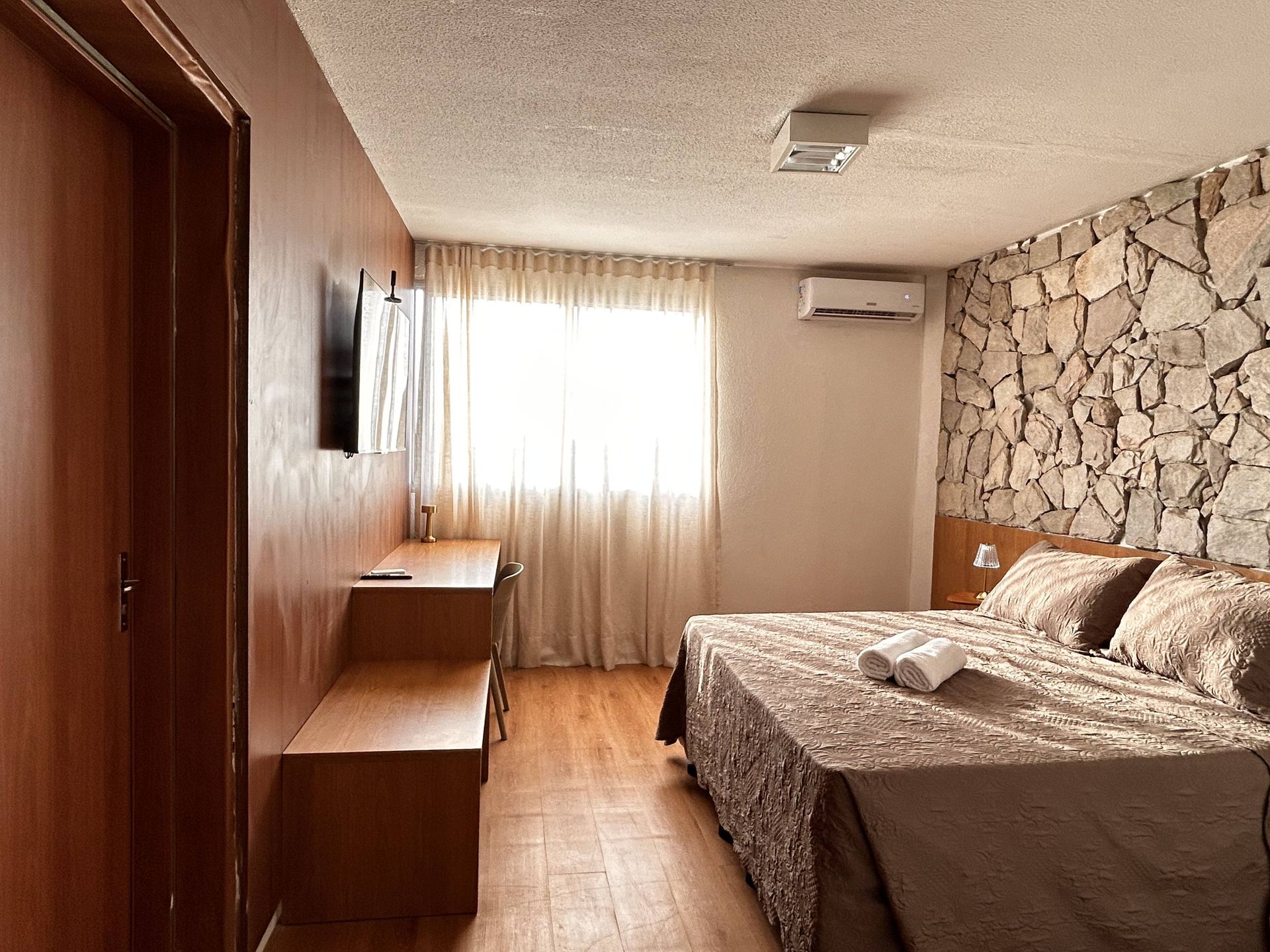 Quarto de hotel com cama, escrivaninha, TV e parede com detalhes em pedra.