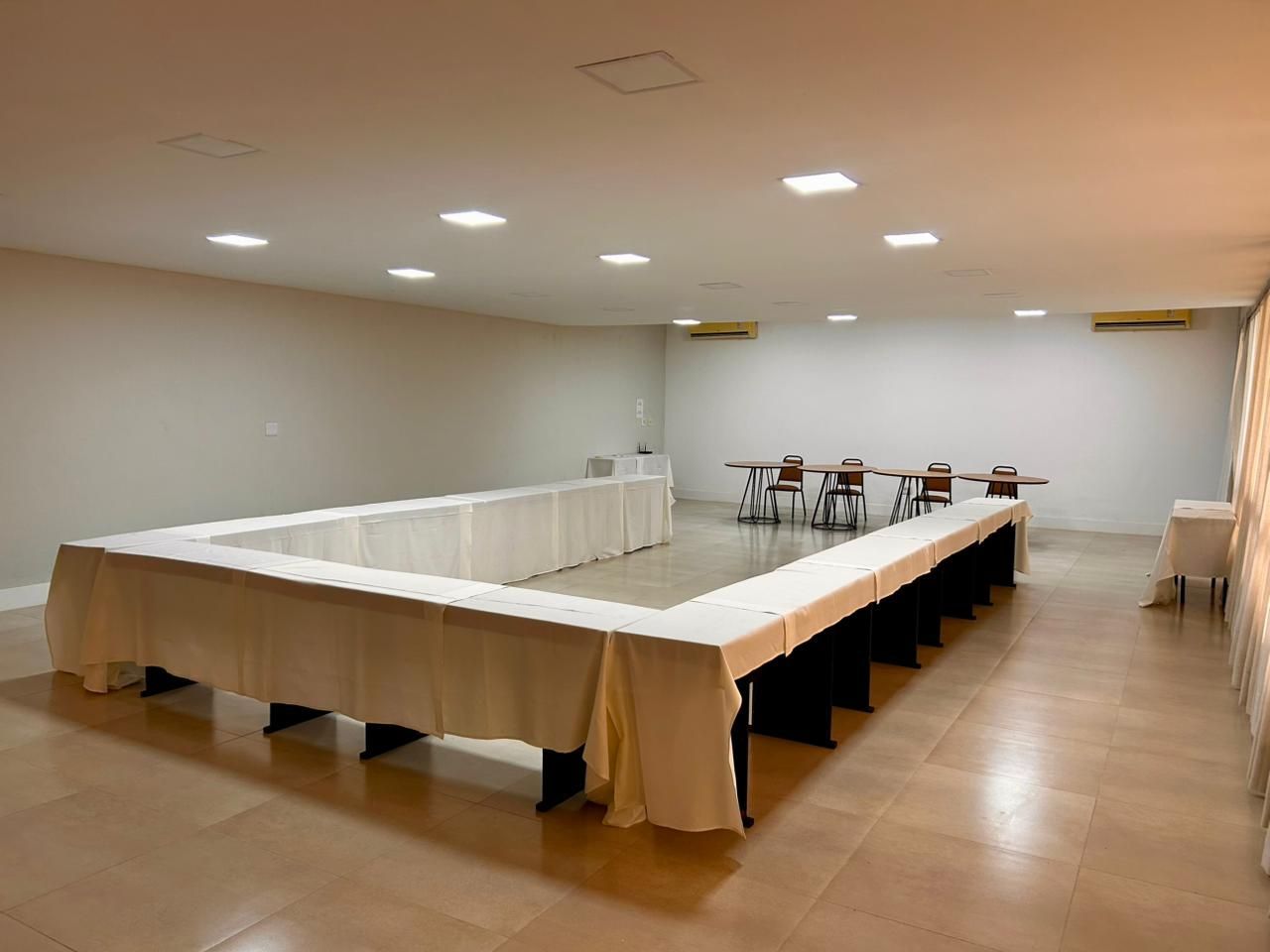 Uma sala de conferências com uma grande mesa em forma de U e cadeiras, piso bege e iluminação brilhante.