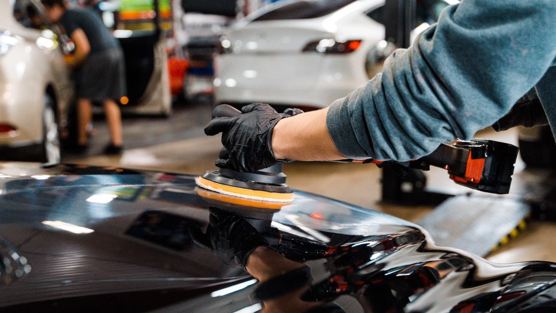 Person polishing a black car in a garage with a power tool while wearing gloves.
