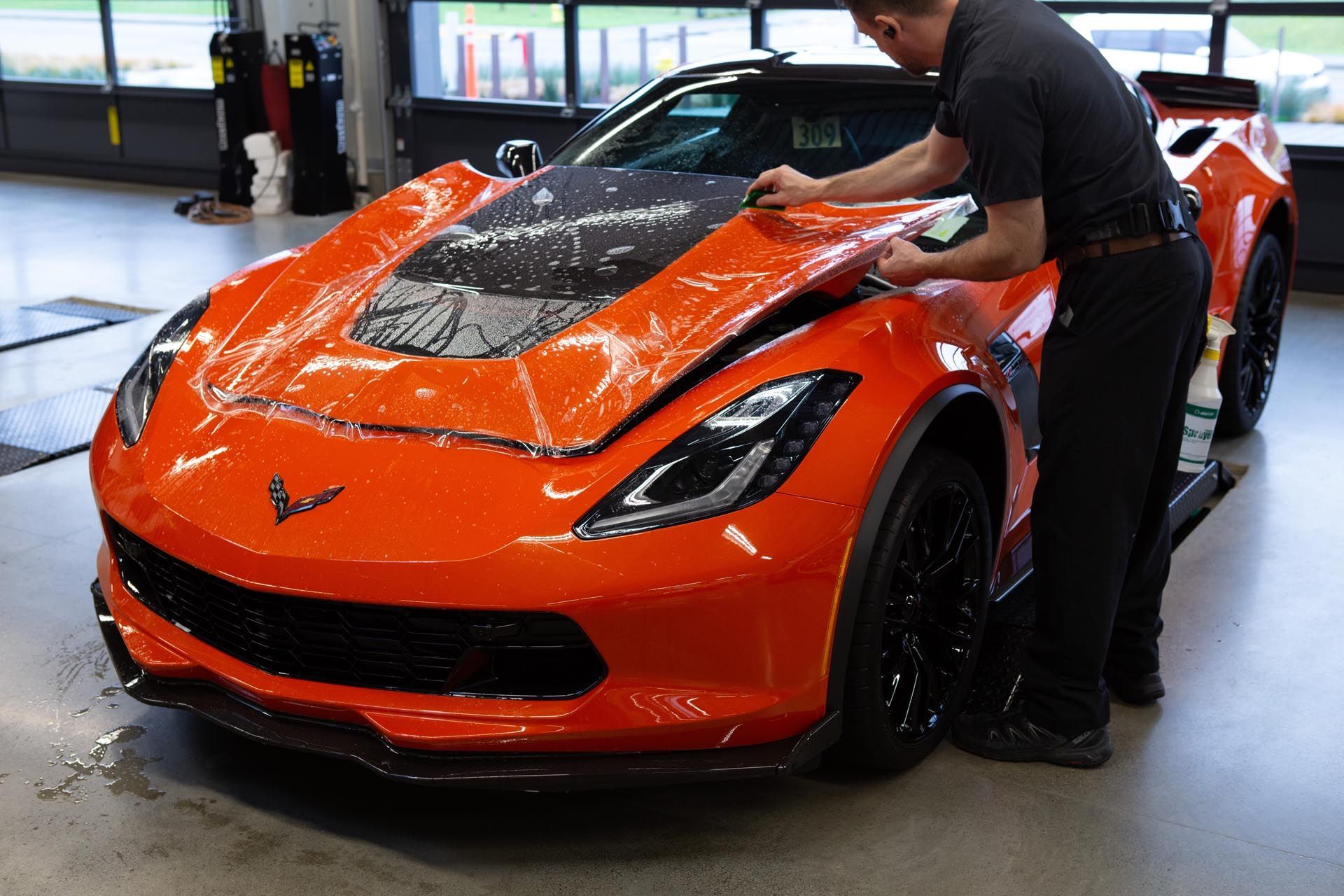 A person applying a clear protective film to the hood of a bright orange sports car inside a garage.
