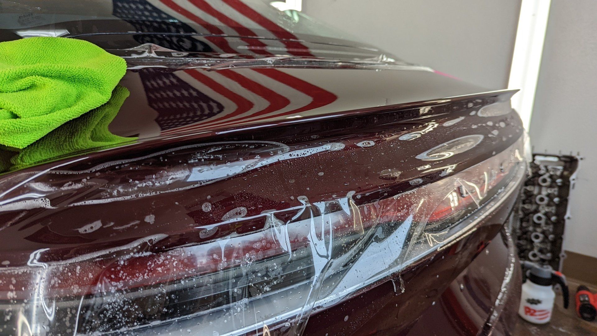 Car's tail light area being wrapped with a protective film, reflection of the American flag visible. Green cloth nearby.