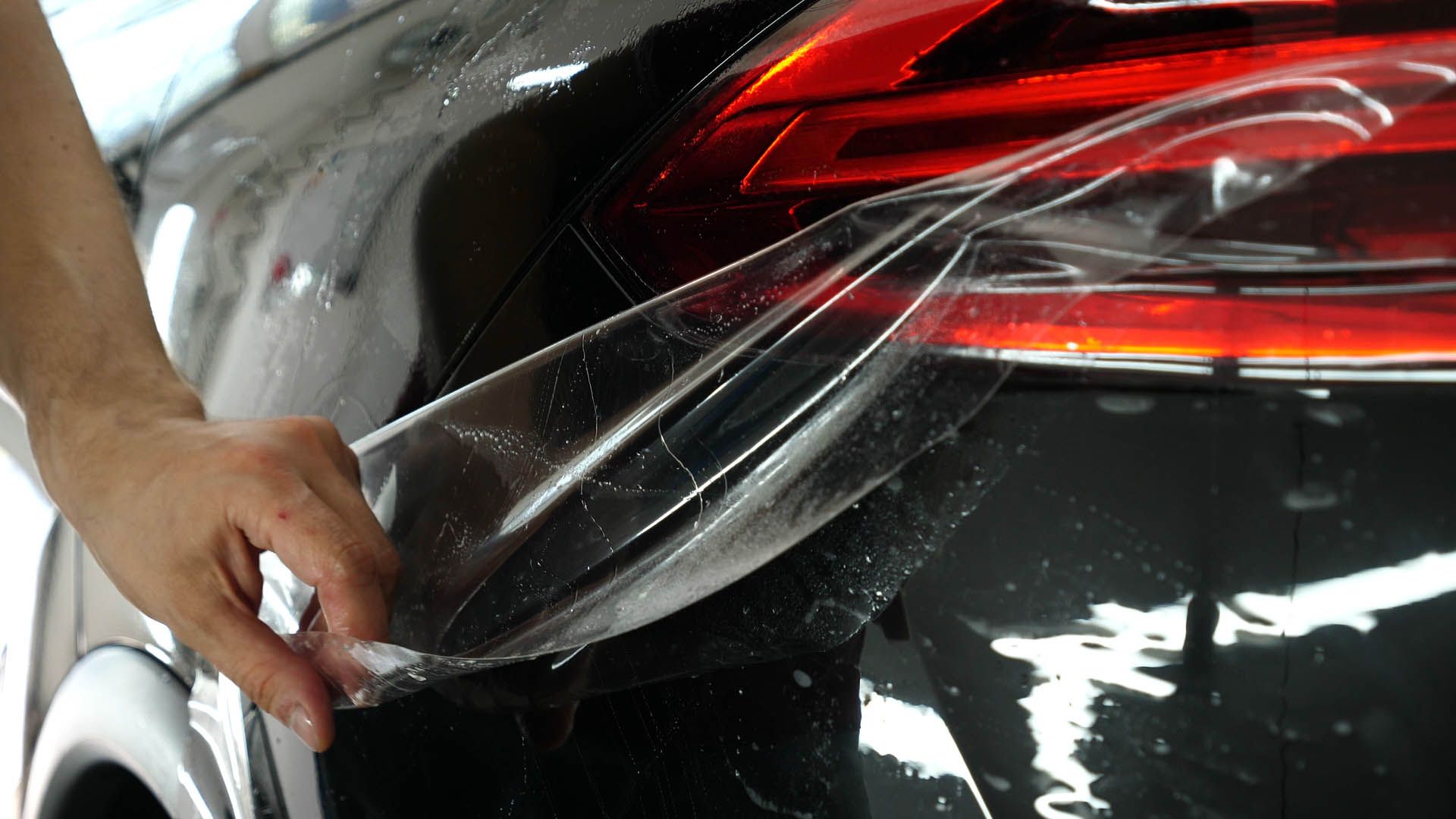 Hand peeling protective film off a black car's taillight.