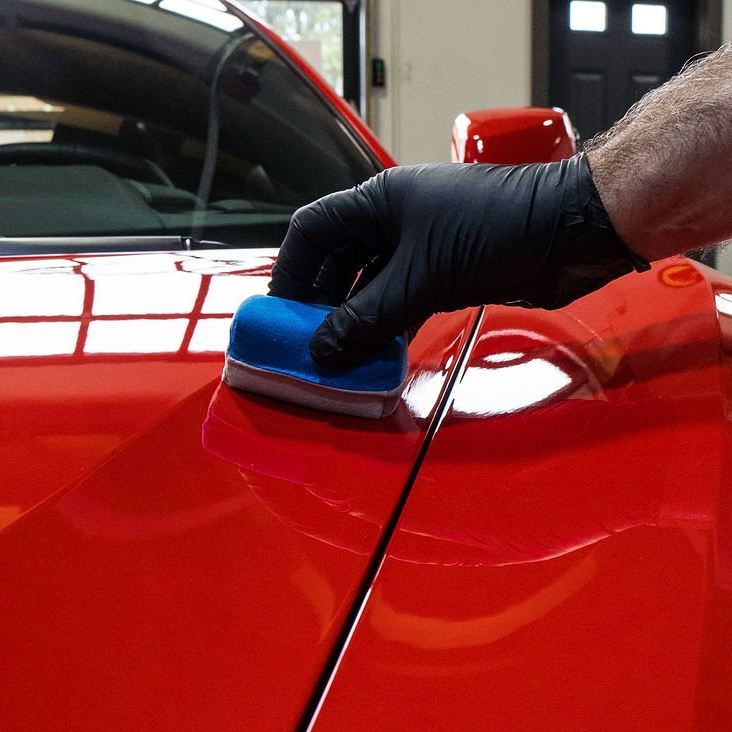 Person wearing a black glove applying a blue applicator to red car paint.