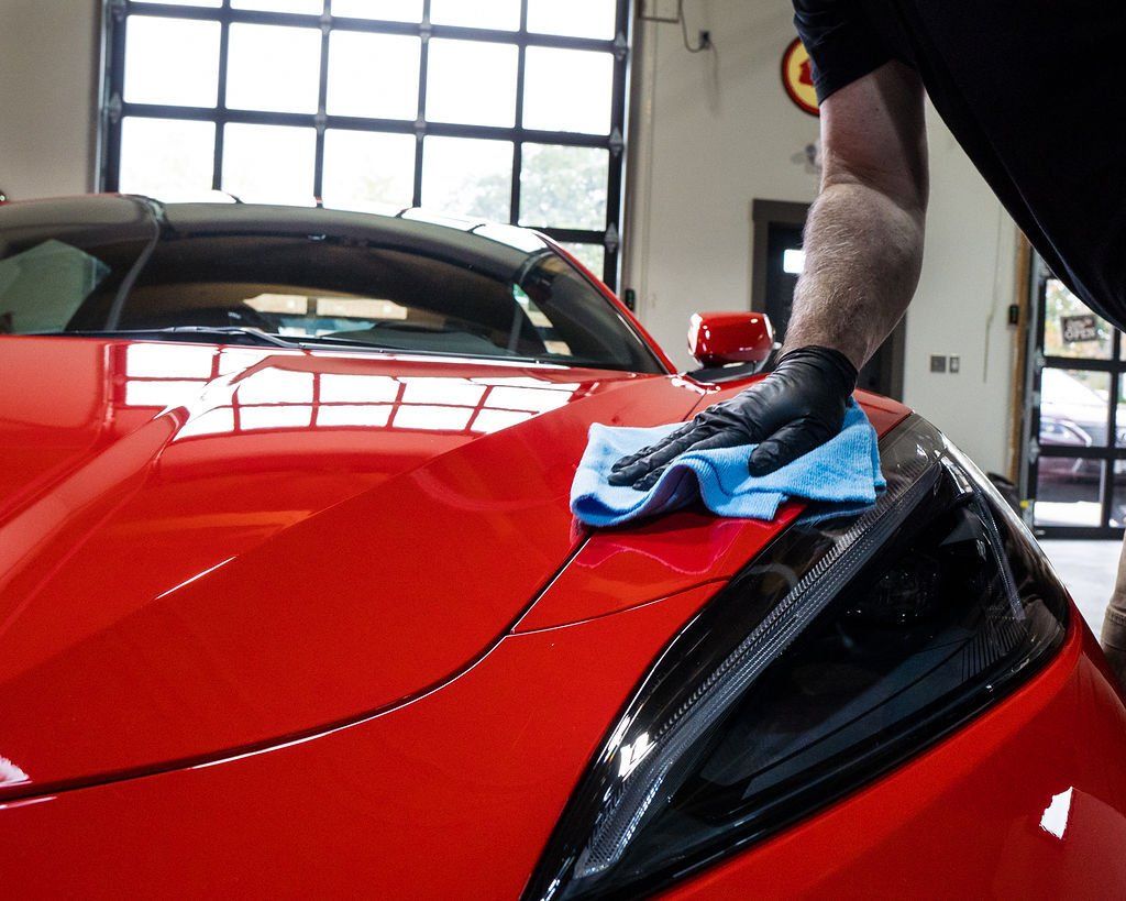 A person in black gloves wiping a bright red car's hood with a blue cloth inside a garage.