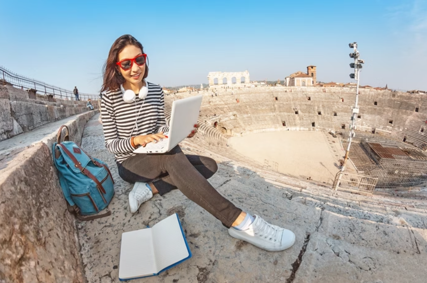 Moça jovem com mochila, caderno e notebook, estudando em um ponto turístico da Europa.