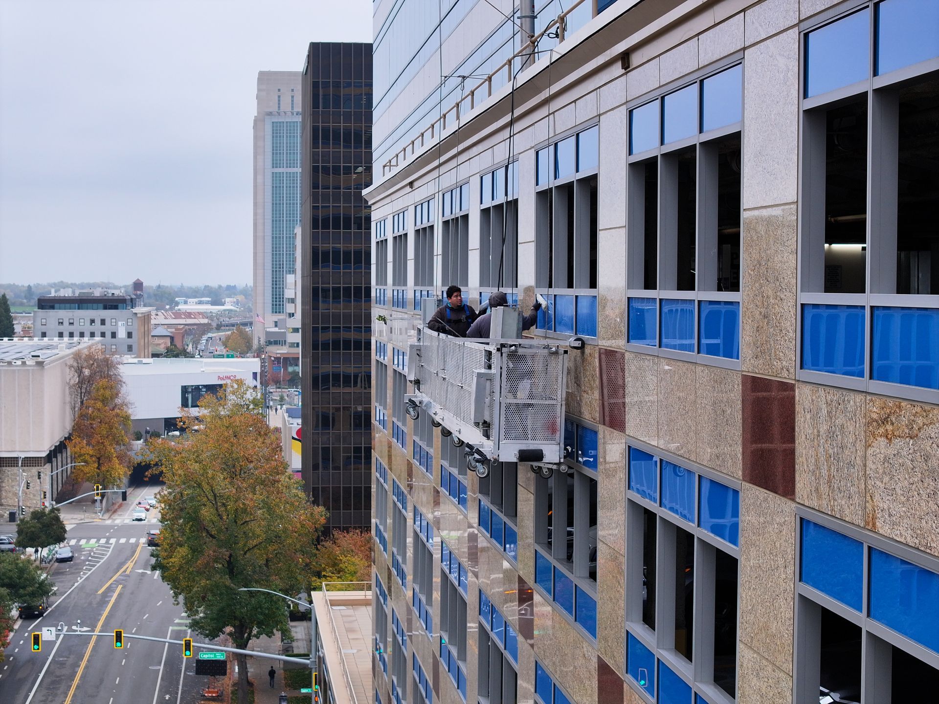Two people on a suspended platform cleaning windows of a skyscraper. City street and buildings in background.