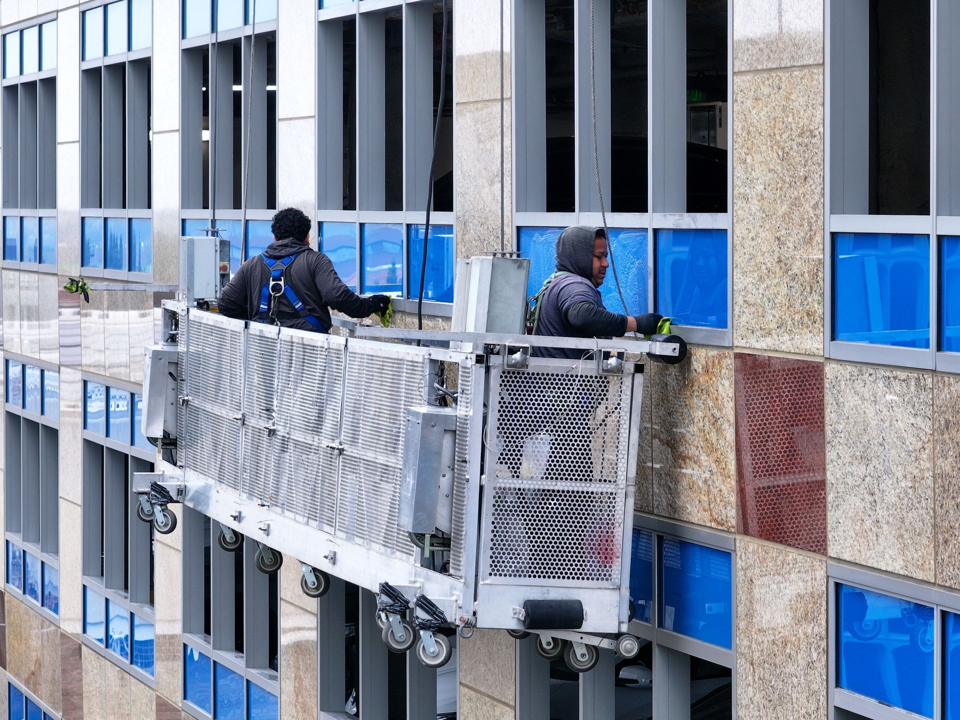 Two construction workers on a suspended platform cleaning exterior windows of a high-rise building.