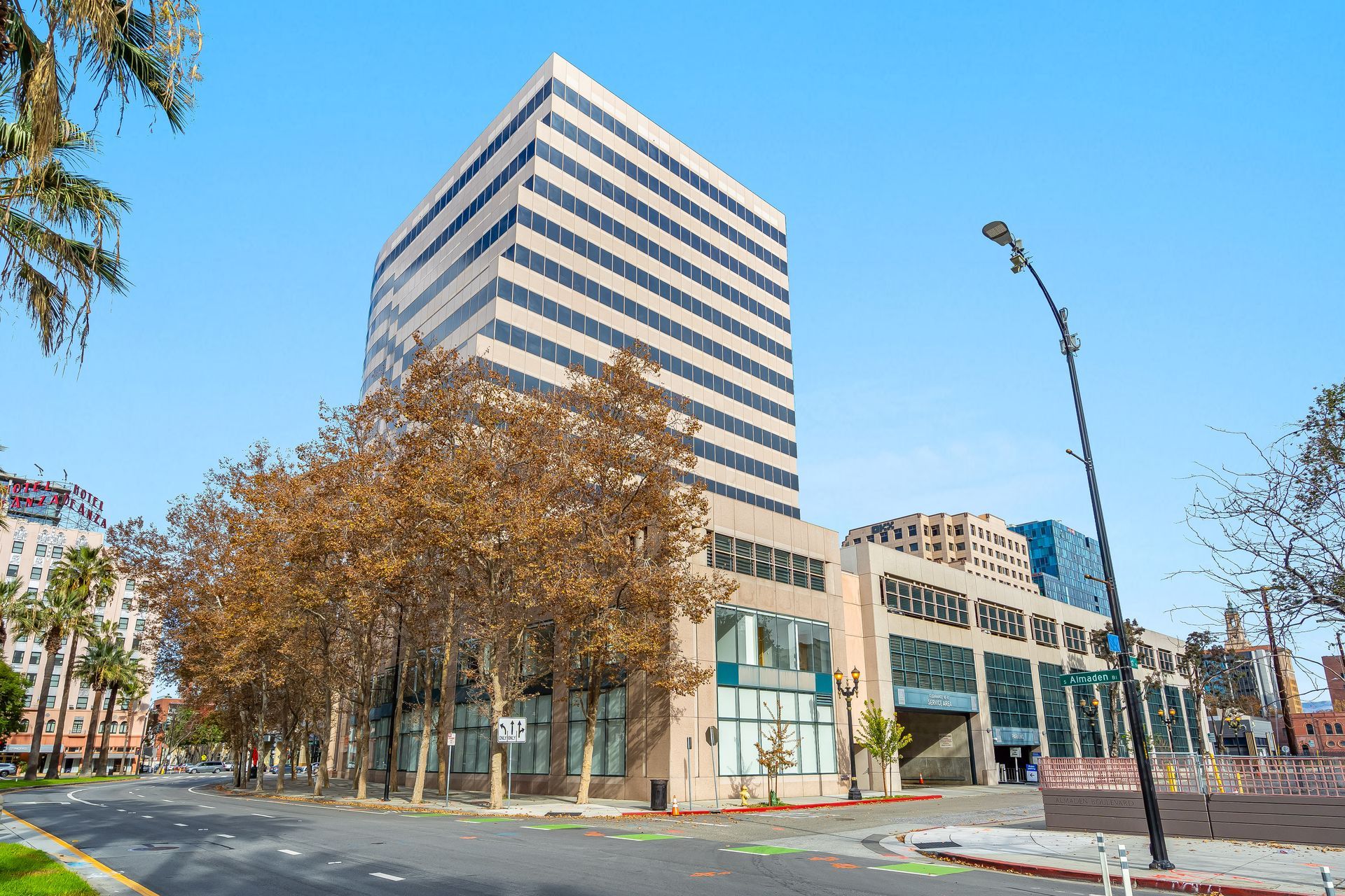 Tall office building with adjacent low-rise structure, trees, and street against a blue sky.