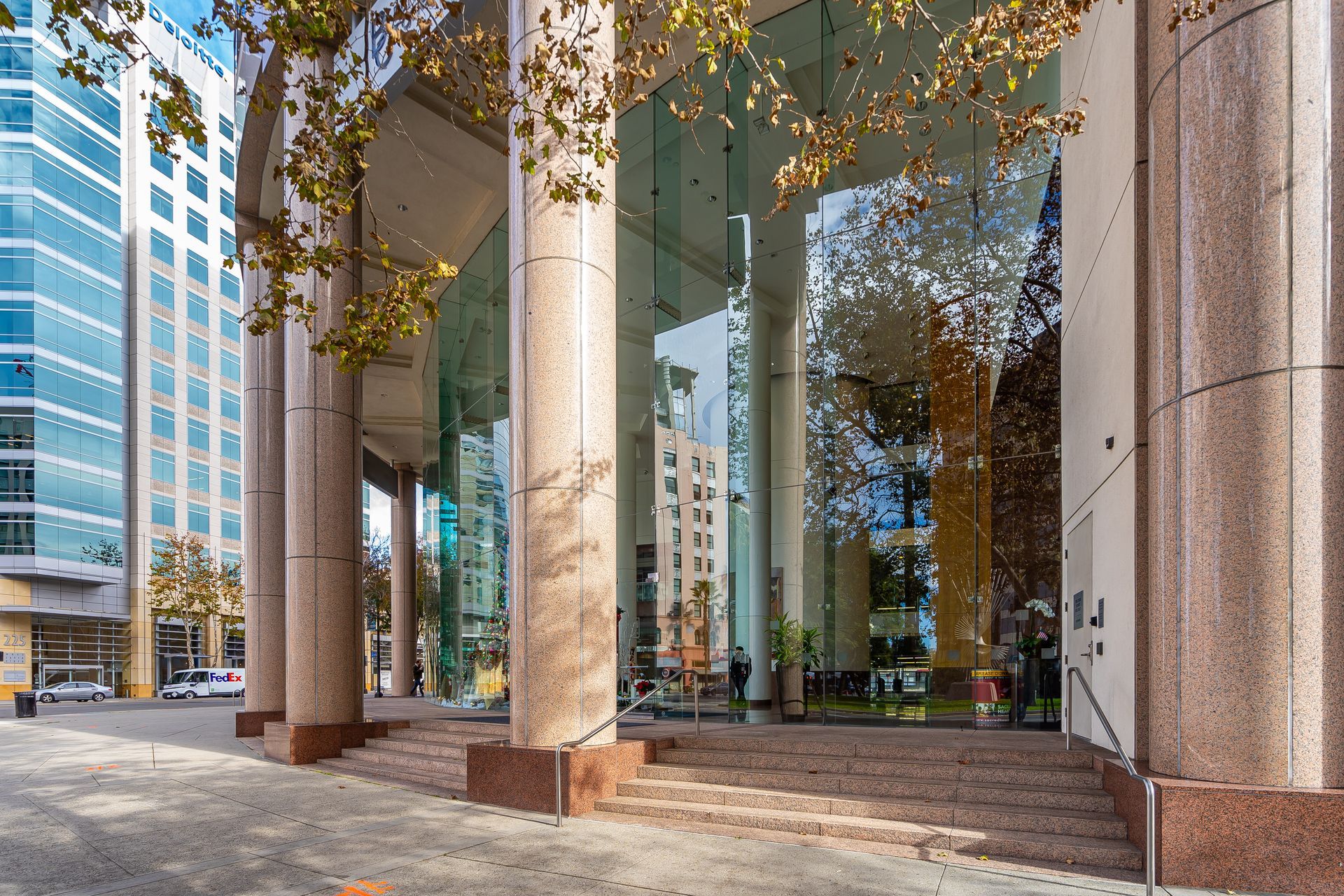 Building entrance with glass walls and large columns; trees and sky reflected.