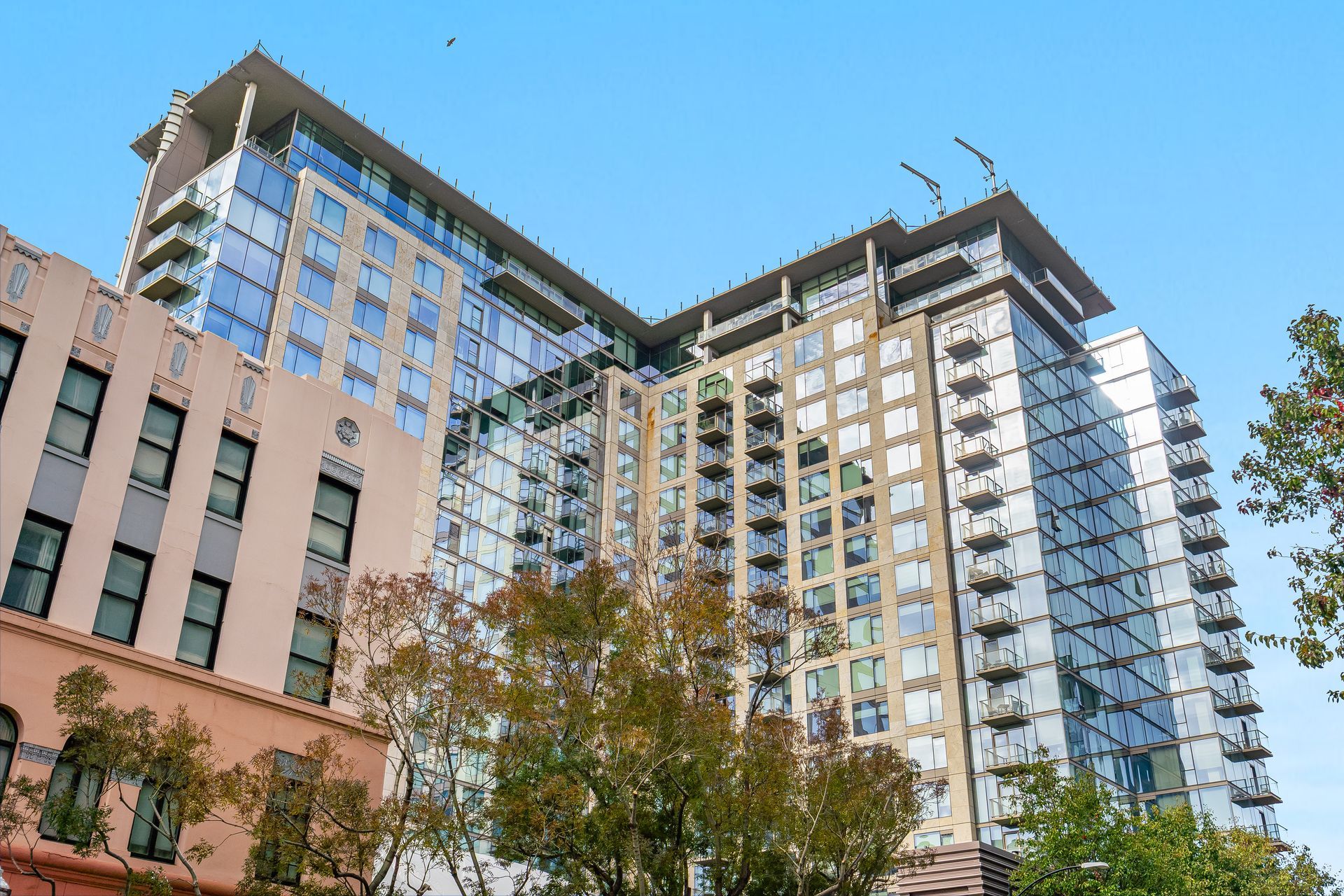 Modern high-rise building with glass and balconies next to an older, light-colored building under a blue sky.