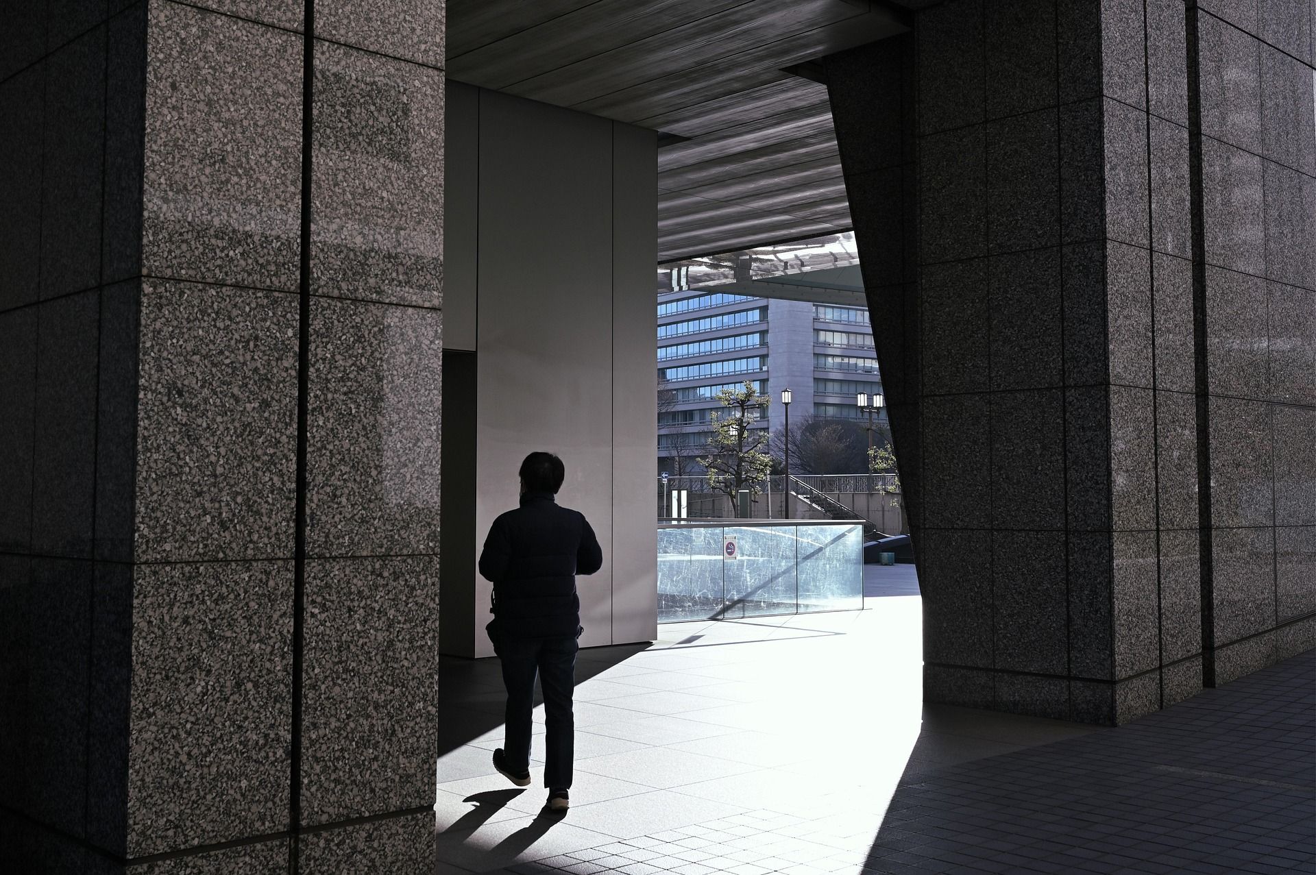Silhouette of a person walking into sunlight between granite columns; city buildings in background.