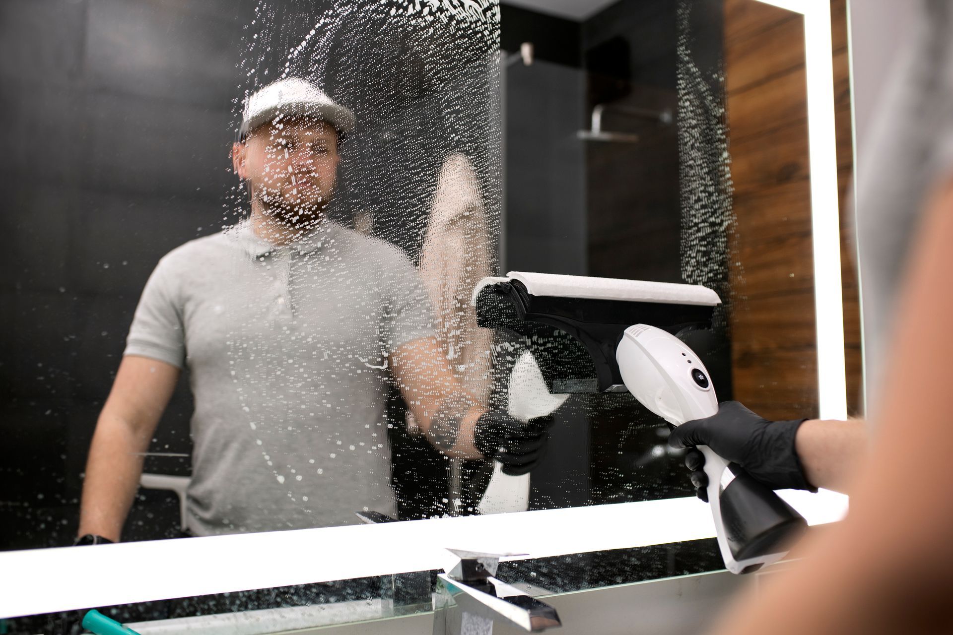 Man cleaning a bathroom mirror with a spray bottle and squeegee.