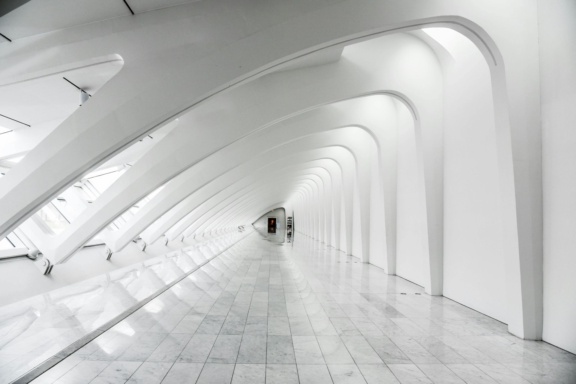 White, arched corridor with a glossy tiled floor. Light streams from above, leading to a distant doorway.