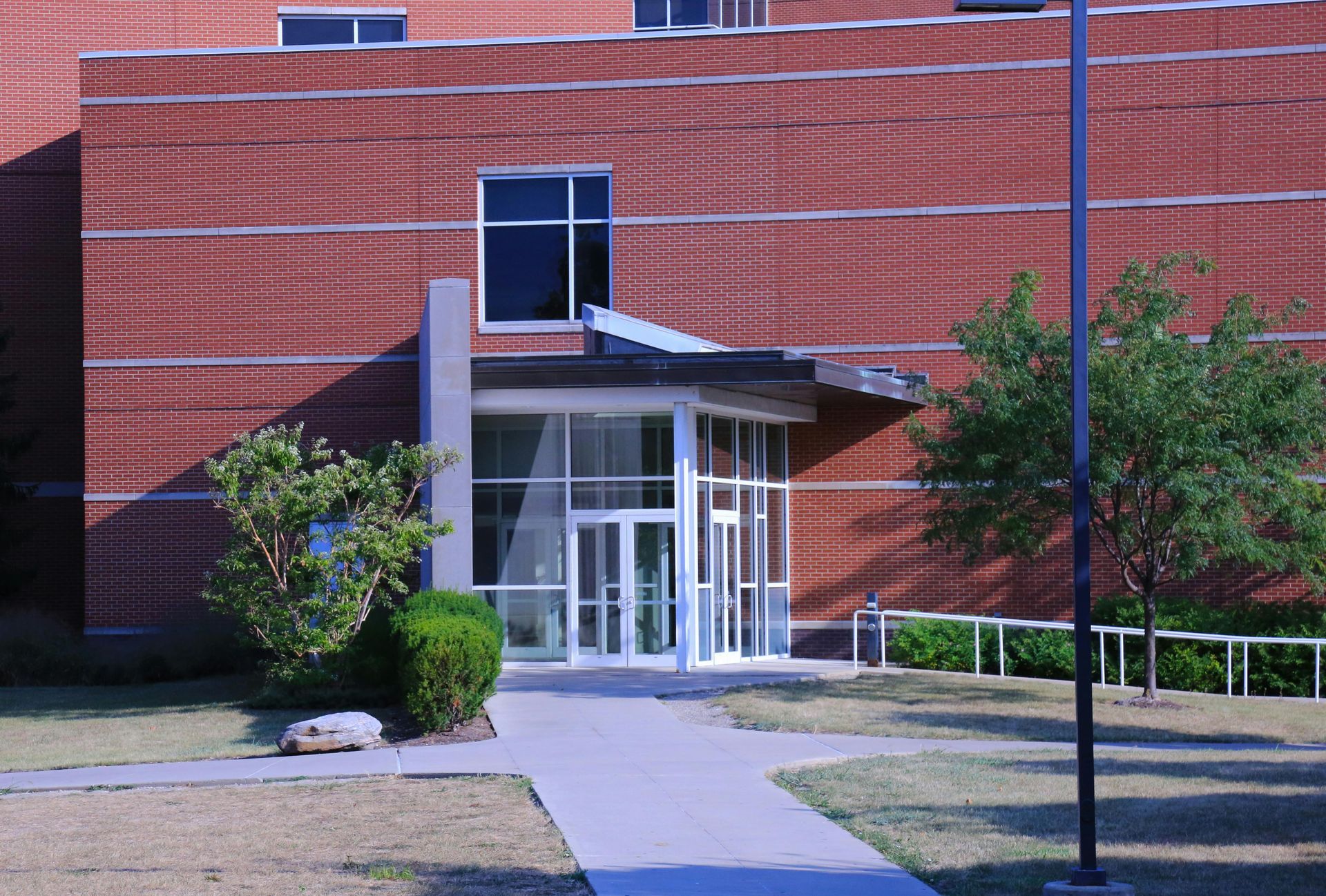 Brick building with glass entrance, concrete pathway, and small trees on sunny day.