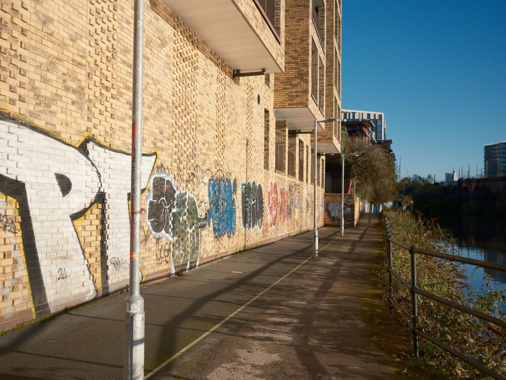 Brick building with graffiti alongside a riverside path, sunny day.
