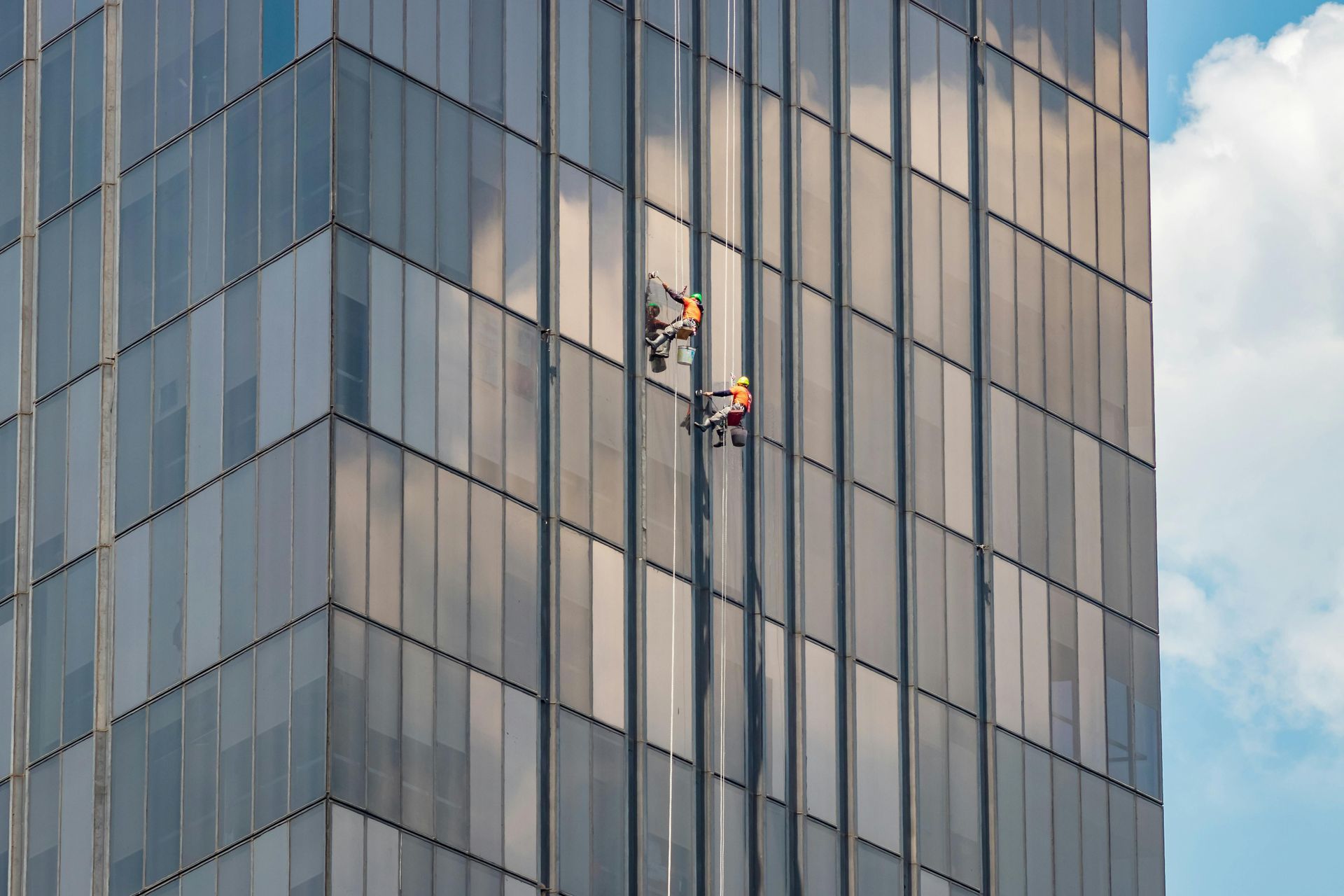 Window washers on a tall, glass-sided building, suspended by ropes. Blue sky and clouds.