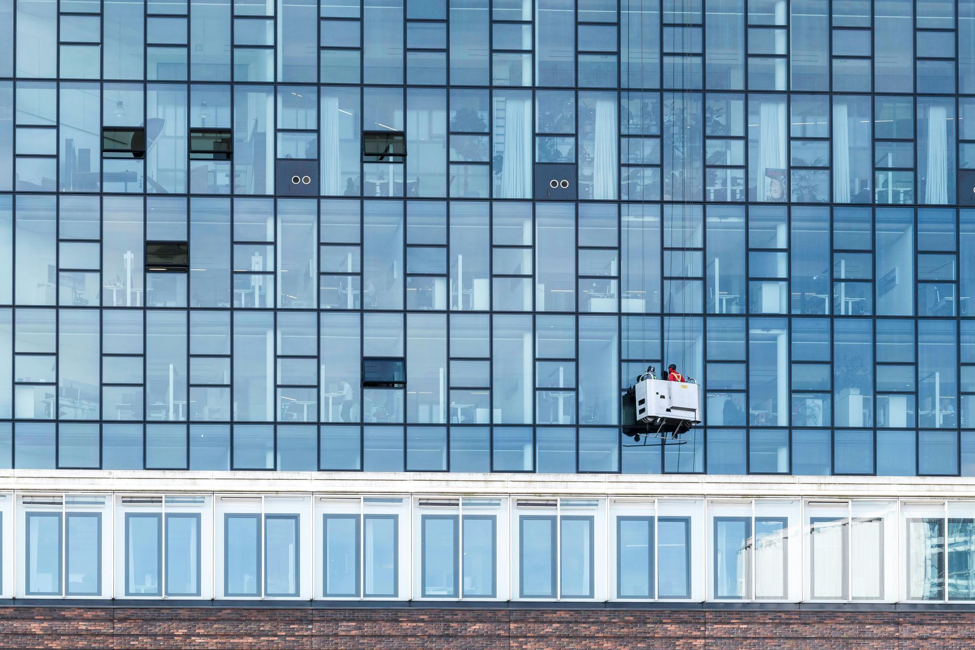Window washer in a suspended platform, cleaning the glass facade of a modern blue office building.