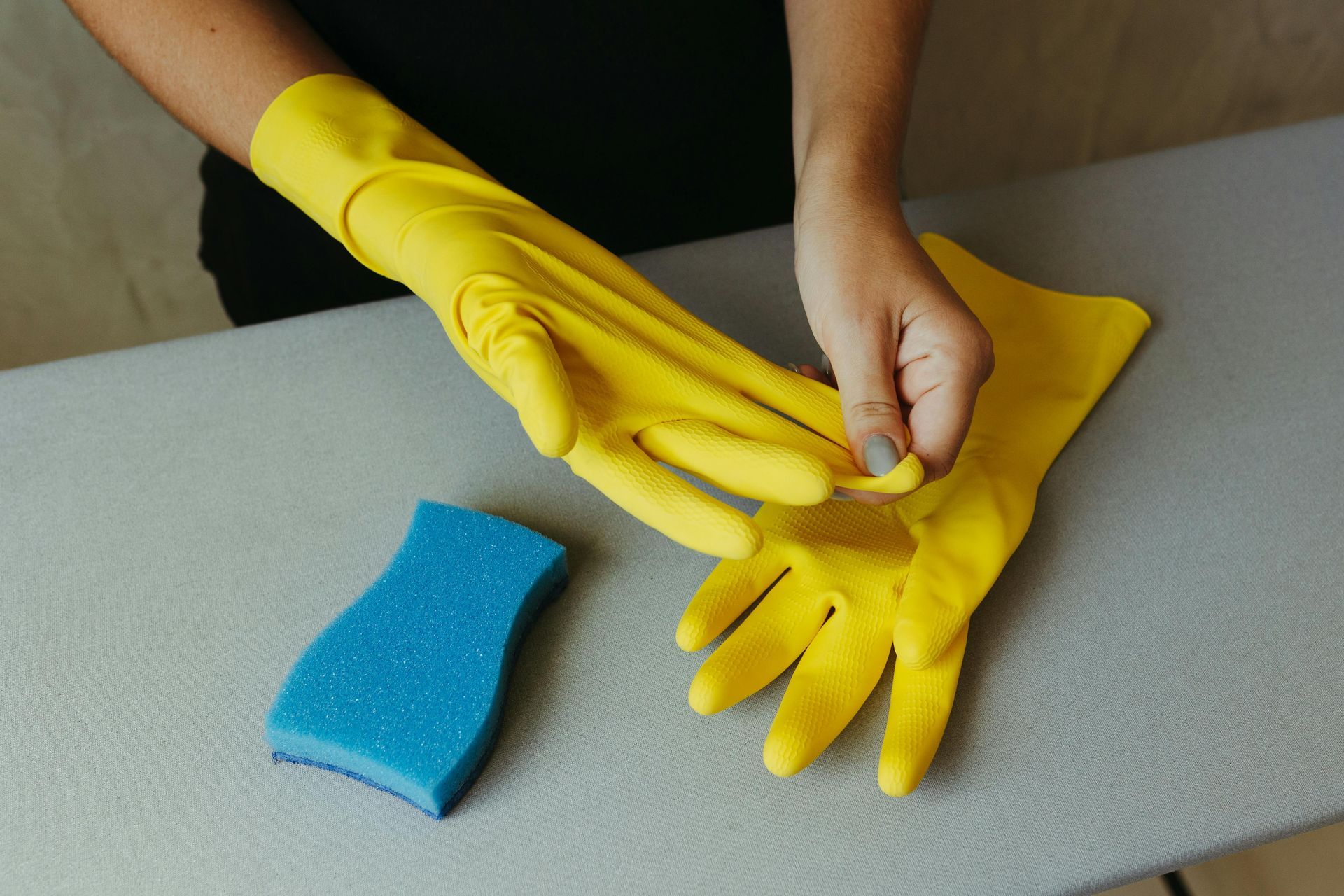 Person putting on yellow rubber gloves next to a blue sponge on a gray surface.