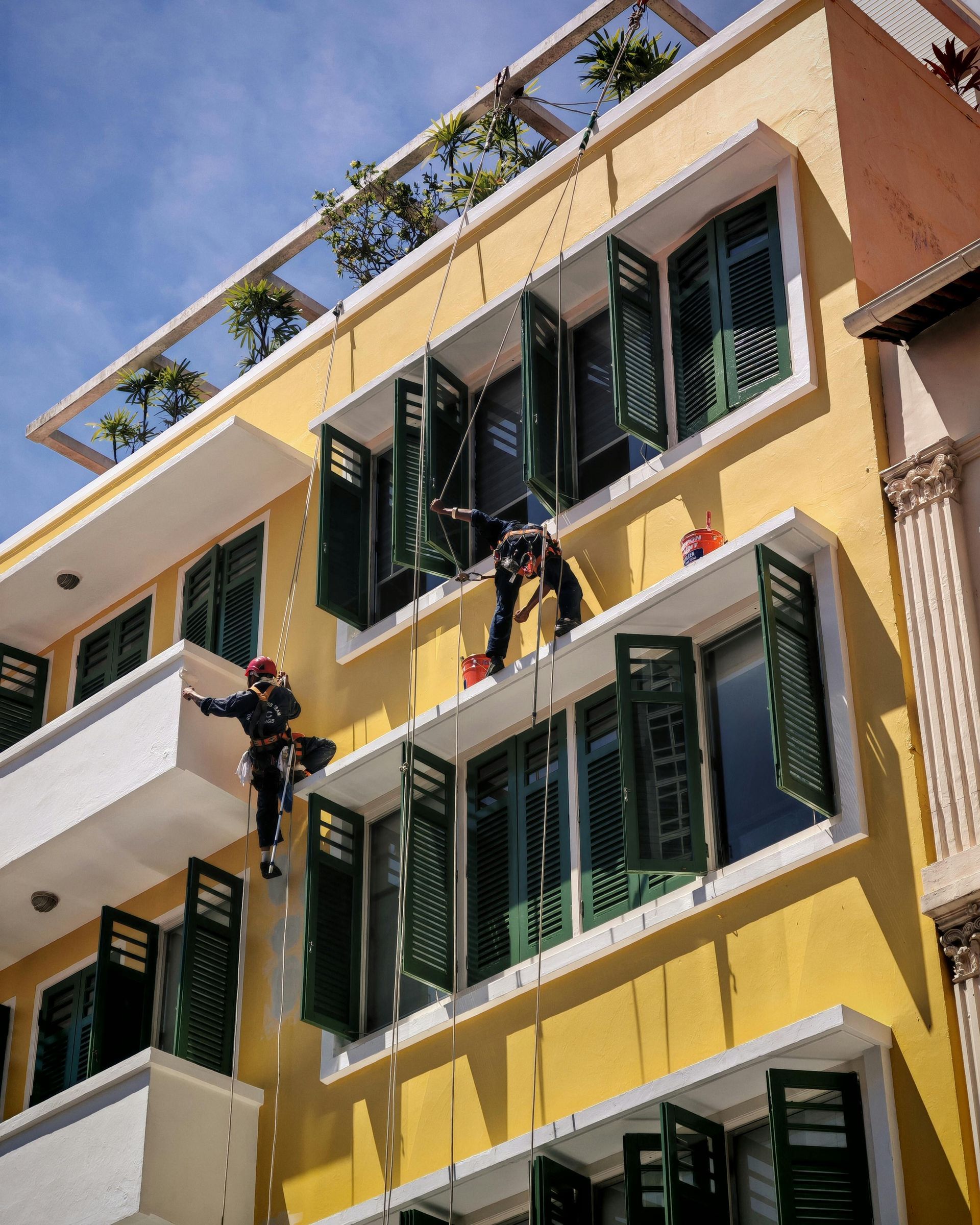 Two window washers on ropes cleaning a yellow building with green shutters and balconies.