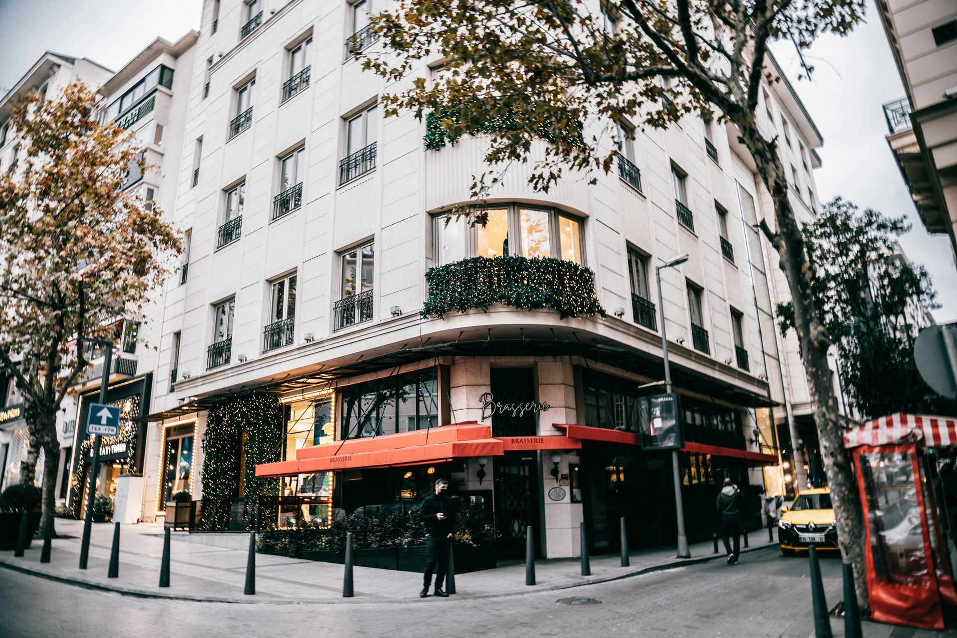 Corner building with a red awning, street scene. A few people stand near the entrance; a taxi is parked nearby.
