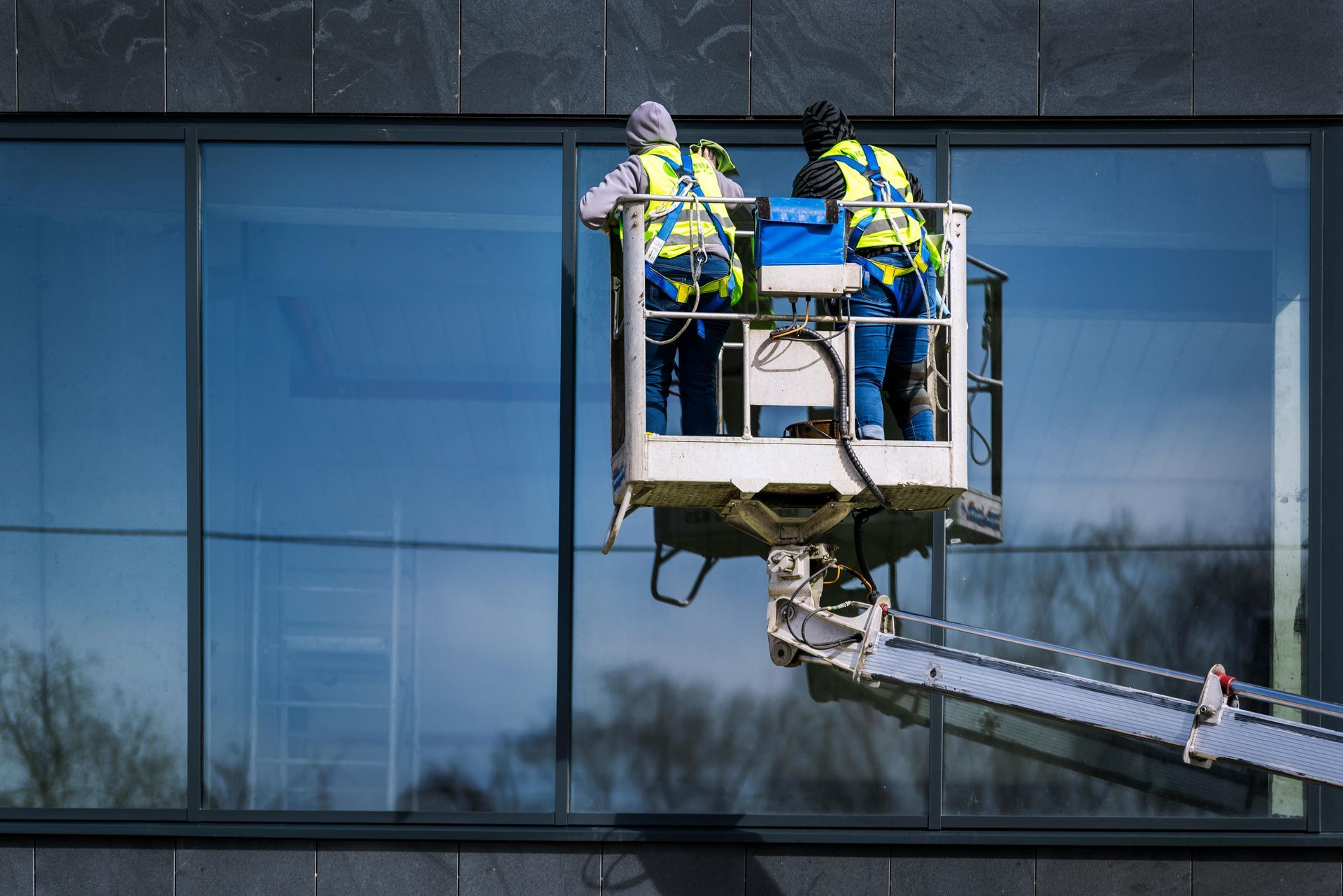 Two workers in safety vests on a lift, cleaning large windows of a modern building.