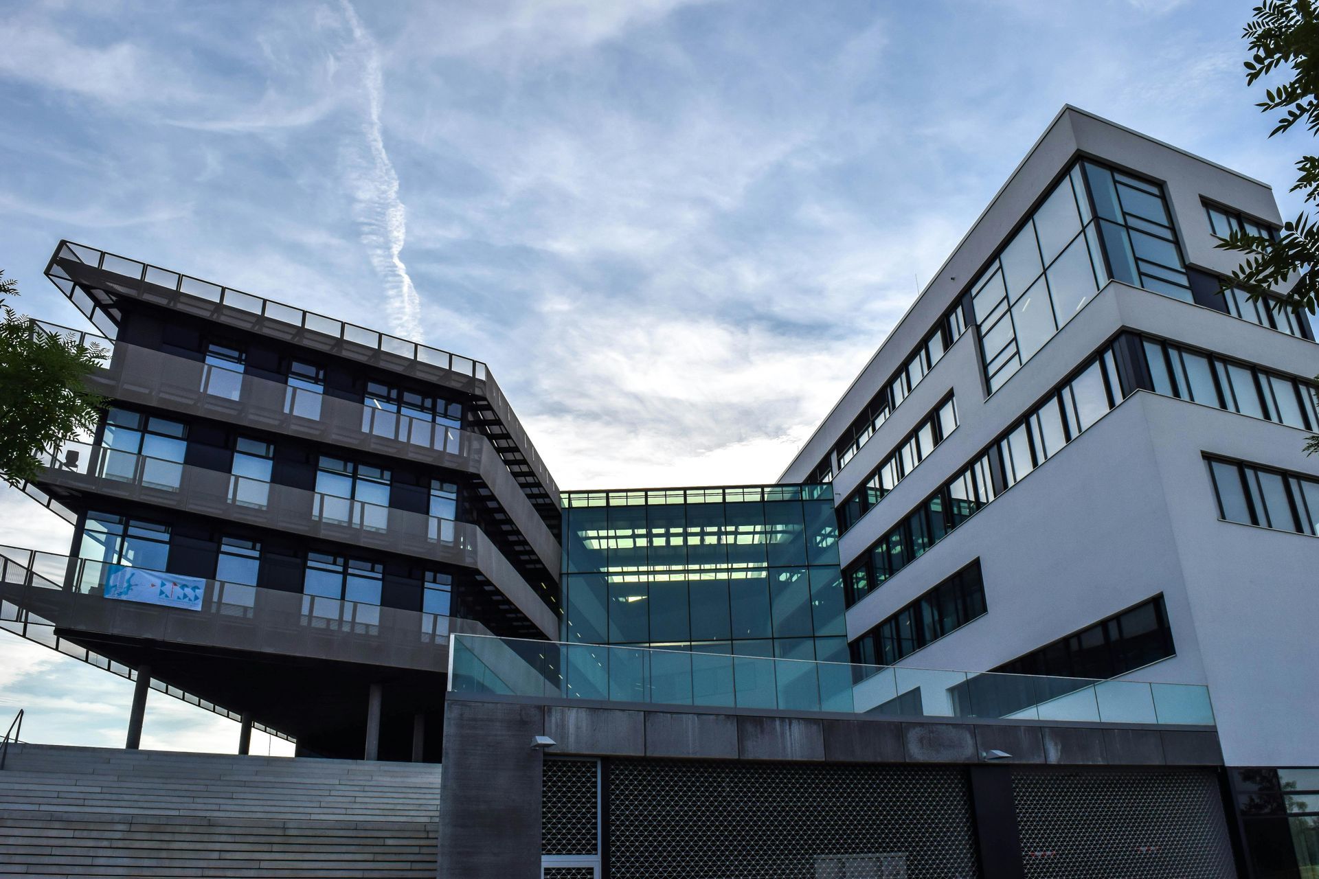 Modern white and gray building with glass bridge, set against cloudy sky.
