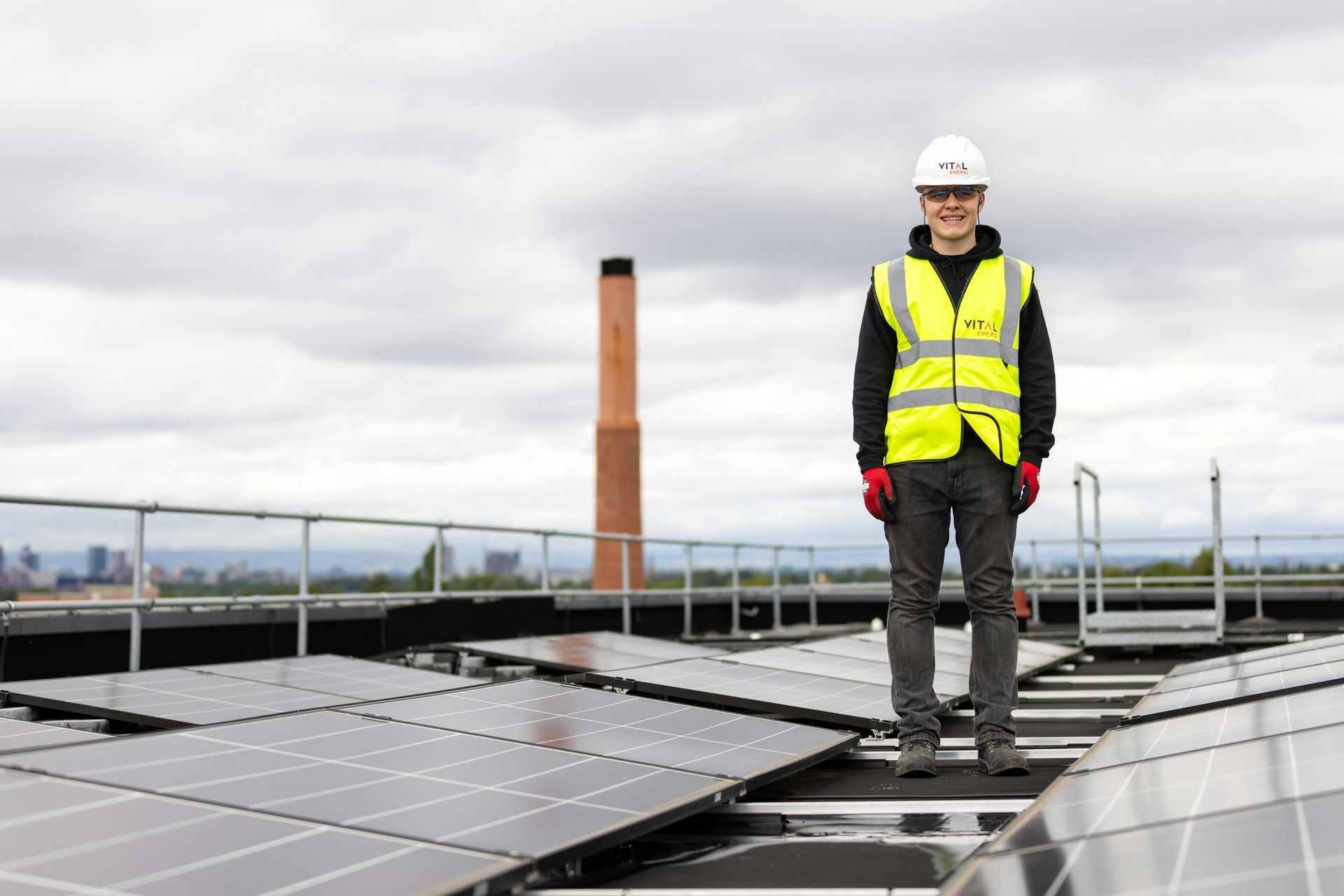 Man in safety gear standing on rooftop solar panels. A chimney and city skyline are visible.