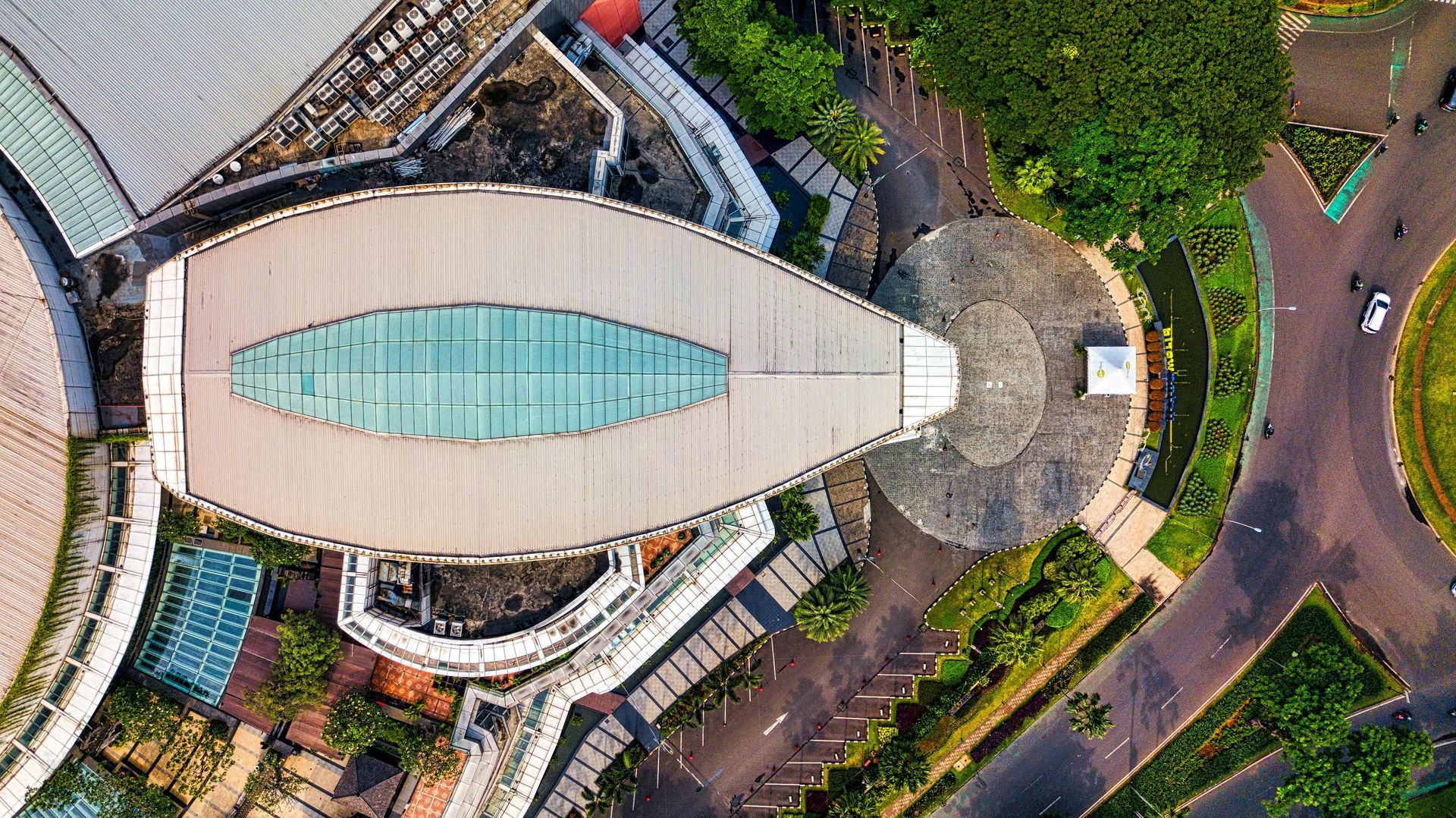 Overhead view of a large, modern building with a distinctive oval-shaped roof and surrounding roads, green spaces.