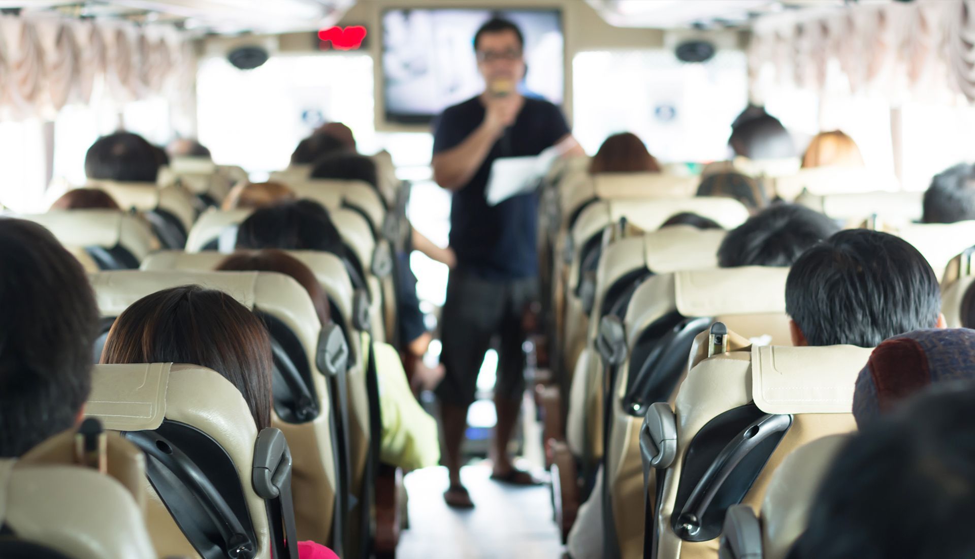 Tour guide speaking in a bus filled with seated passengers; interior shot.