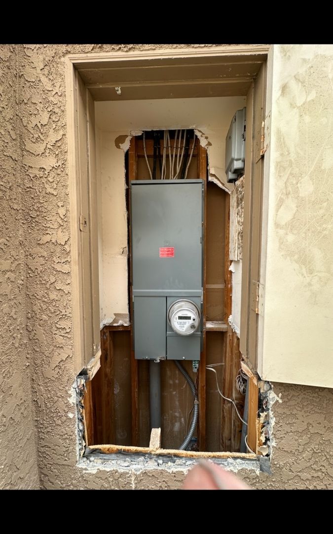 Electrical box recessed in a wall. Gray metal box, meter, exposed wiring, and wooden framing.