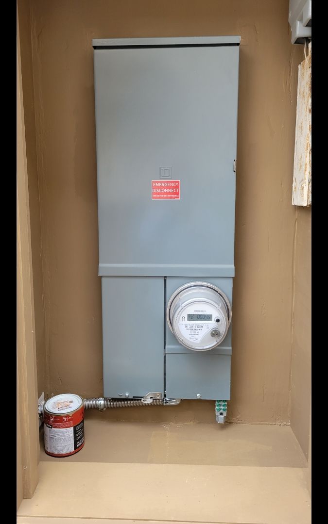 Gray electrical panel with meter, mounted on a tan wall. Grounding wire and paint can are visible.