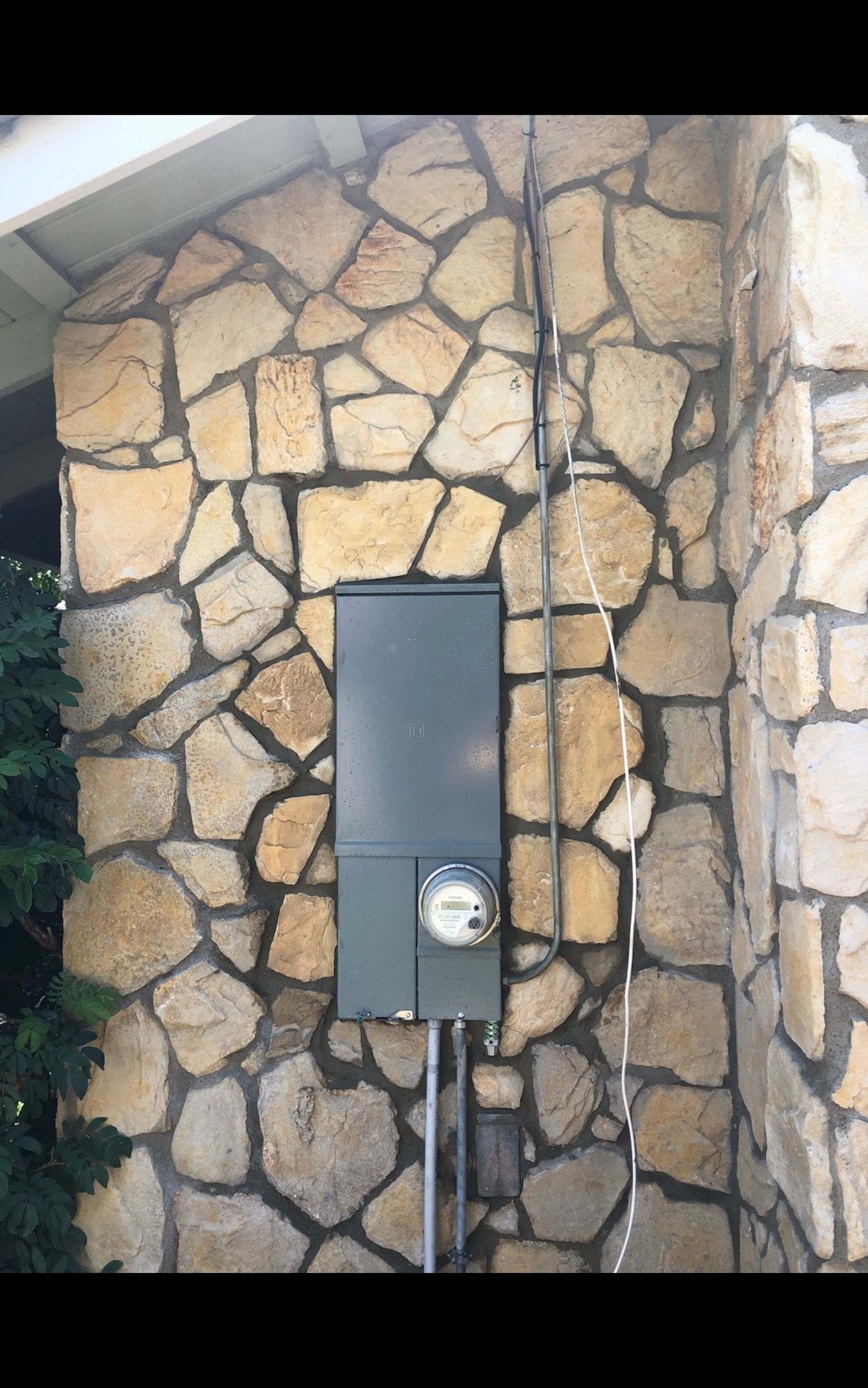 Electric meter and service panel on a stone wall. Gray panel and meter with conduit.