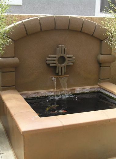 Fountain with a New Mexico Zia sun symbol, water flowing into a dark pond, tan stucco walls.