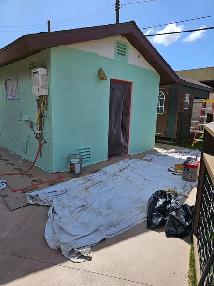Small green building being painted. White tarp and supplies are on the ground.