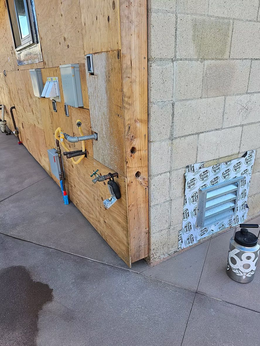 Exterior building corner with plywood siding, concrete blocks, and electrical boxes.
