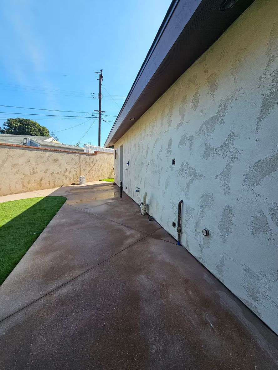 Backyard with a concrete patio, green grass, and a stucco wall under a blue sky.