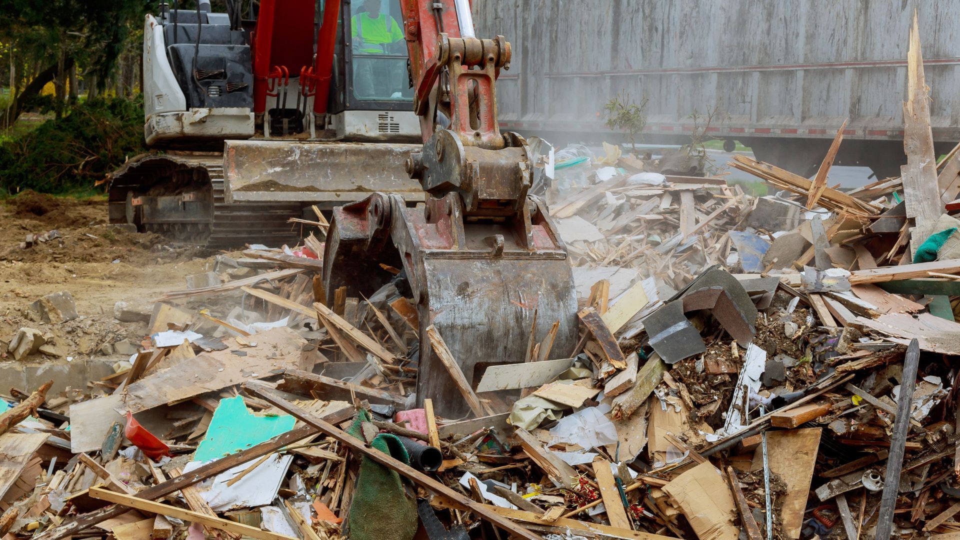 Excavator demolishing a building, scooping debris into a truck, outdoors.