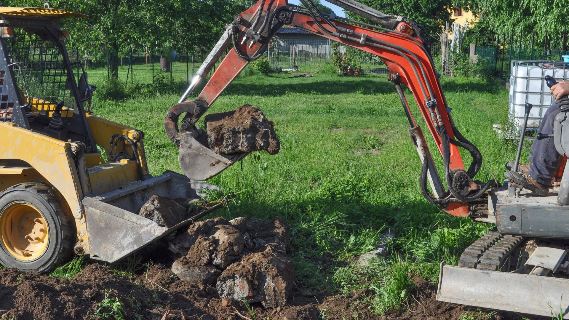 Mini excavator dumping dirt into a front loader bucket in a grassy area.