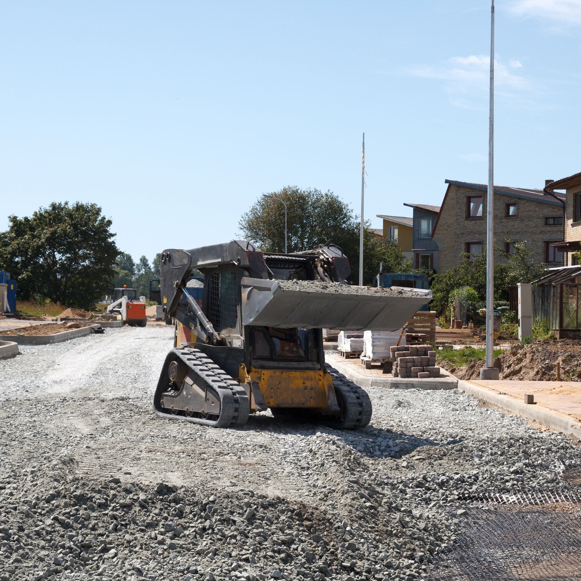 Construction site: Yellow skid steer spreading gravel on a road. Houses and trees in background.