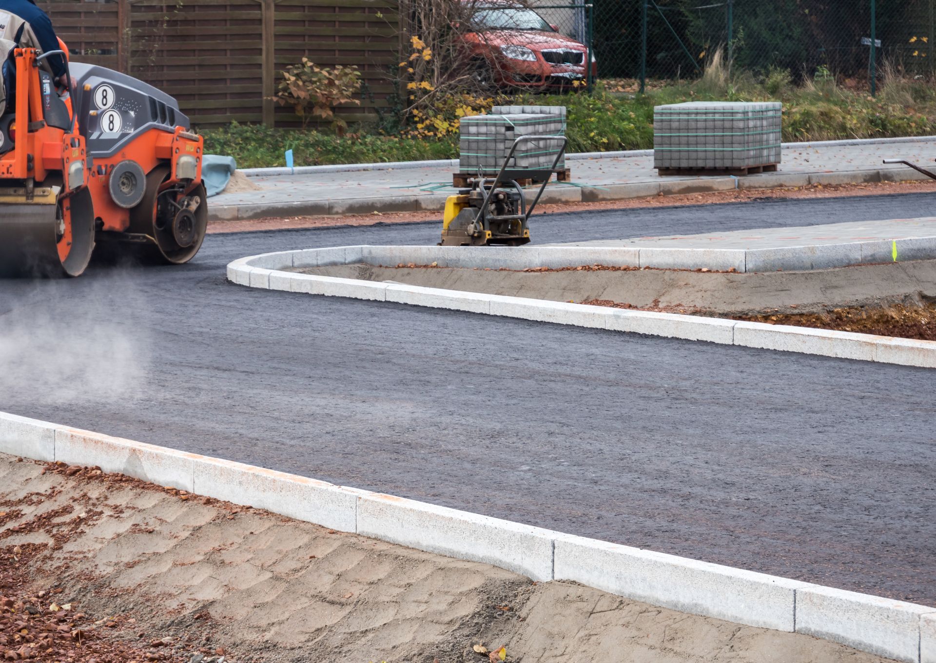 Road construction: Asphalt being laid with a roller and compactor; concrete curb in foreground, overcast day.