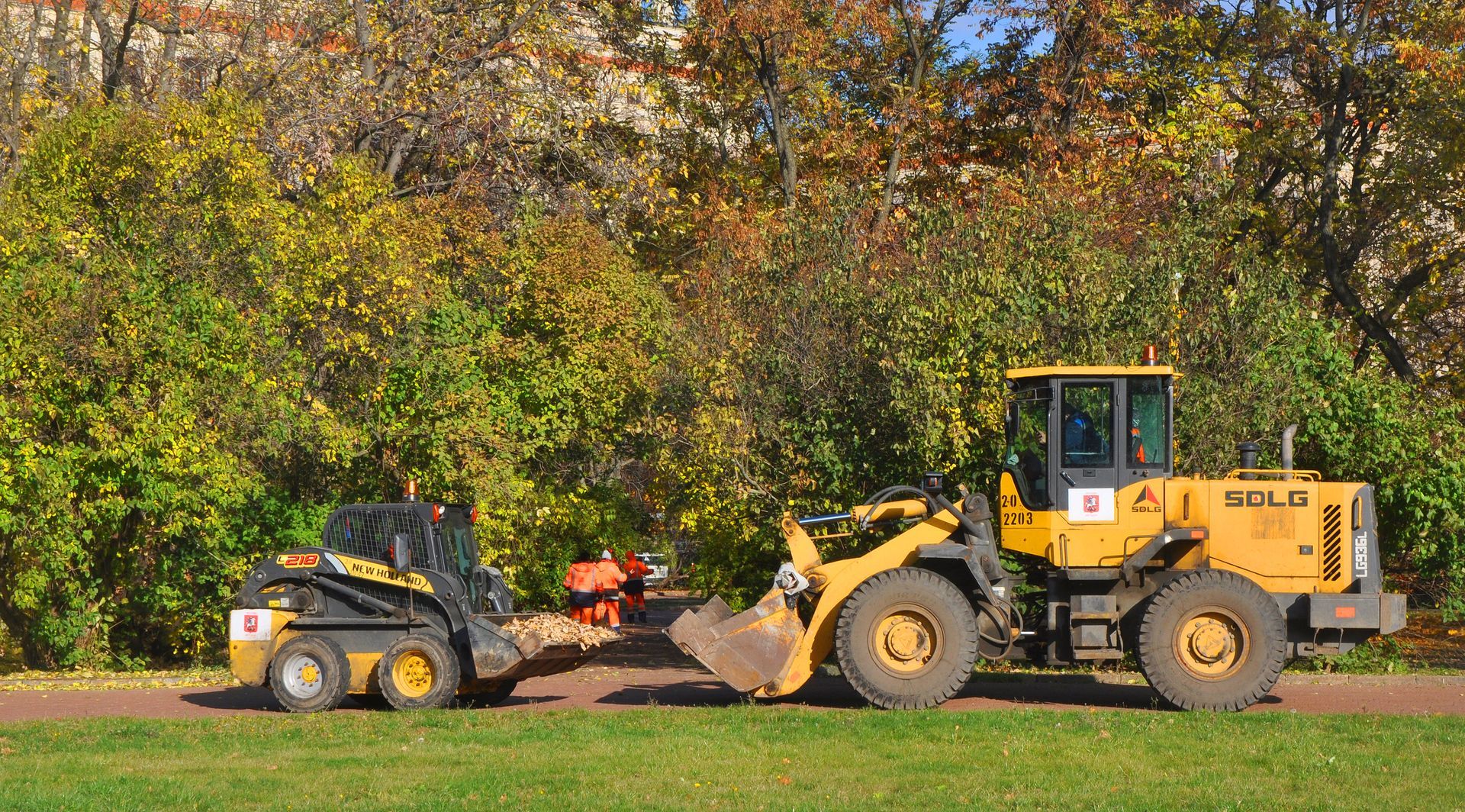 Two yellow construction vehicles on grass path. Trees in background with autumn leaves.