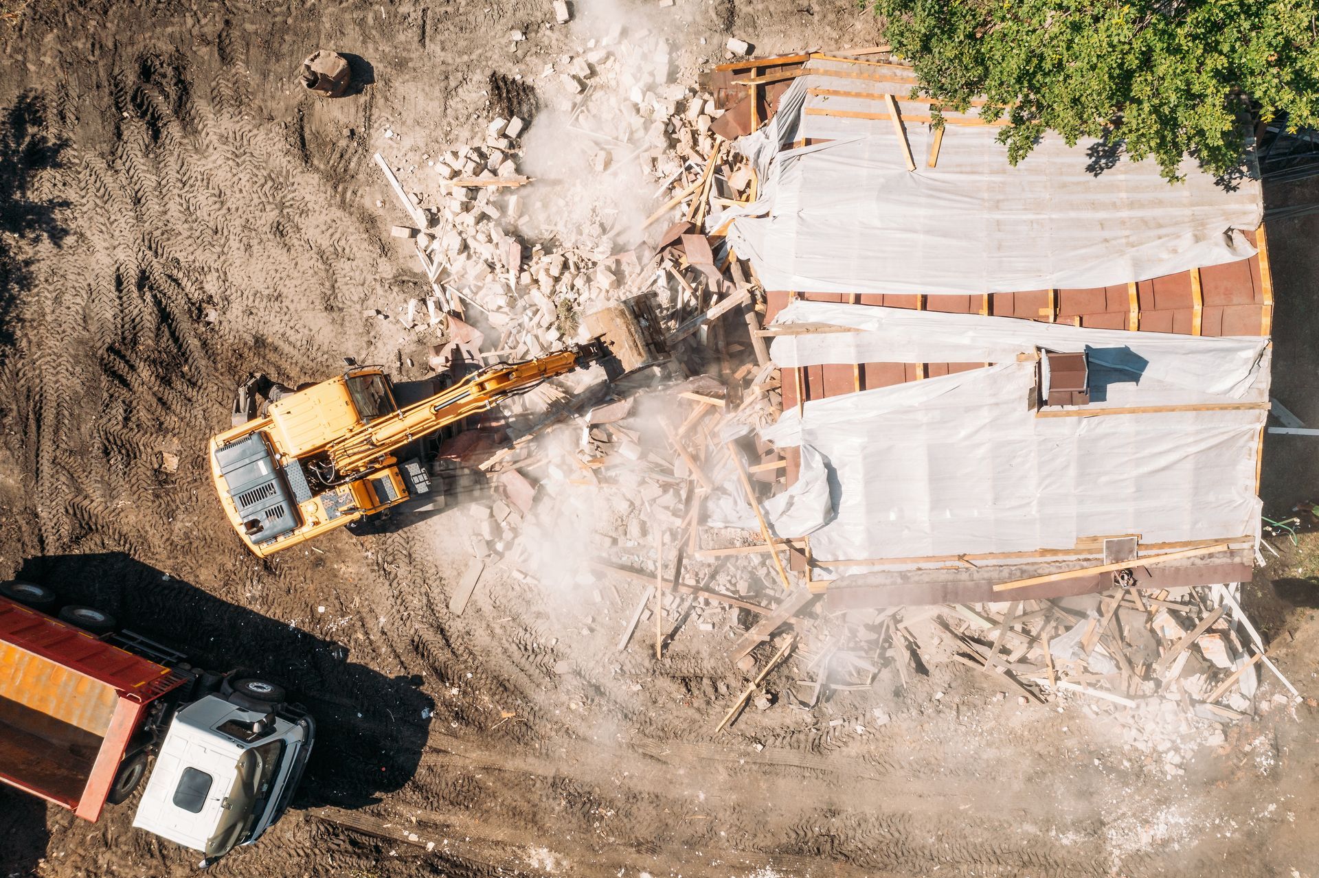 Aerial view: Yellow excavator demolishing a building; debris, a truck, and trees are visible.